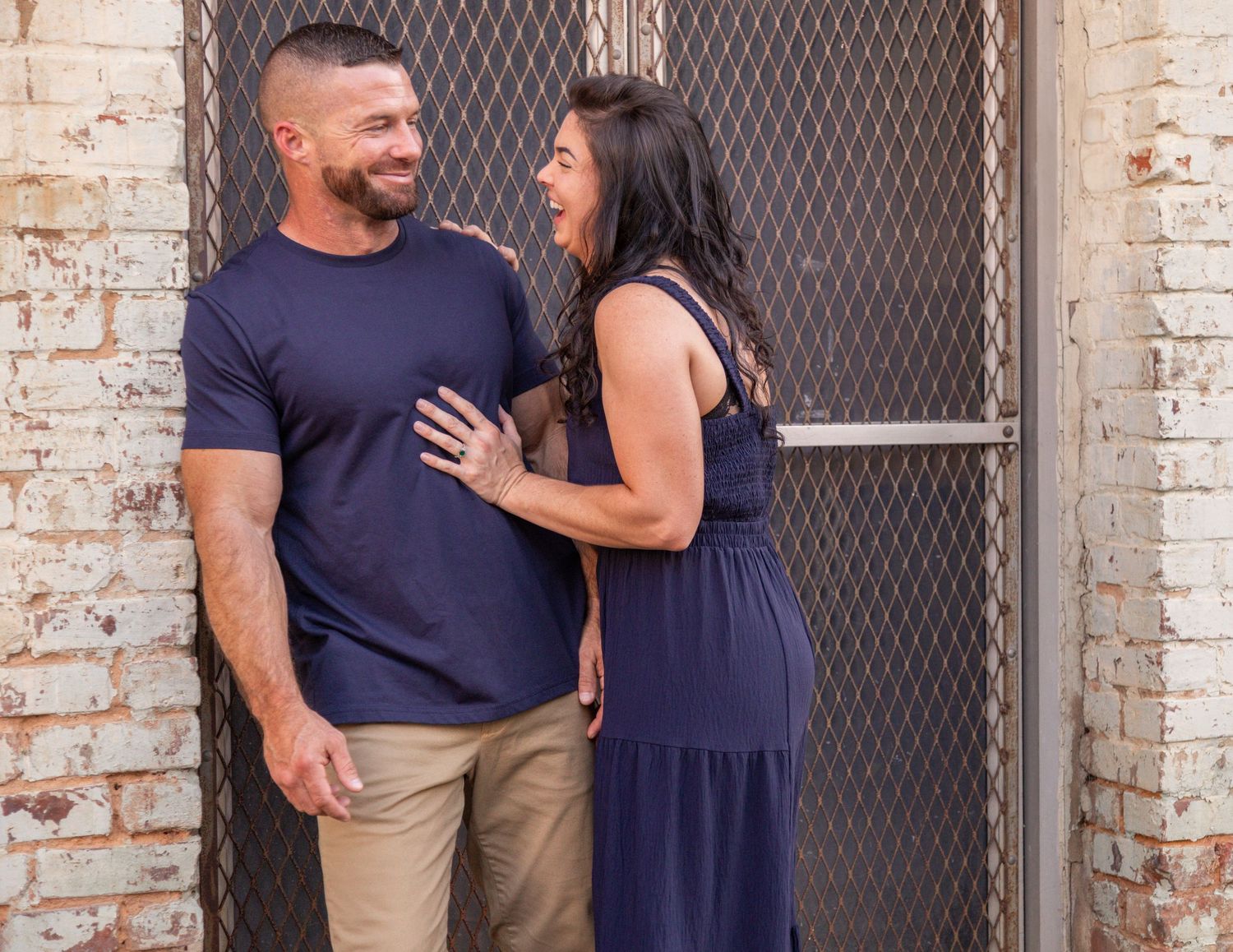A couple shares a playful moment while standing against a rustic brick wall.