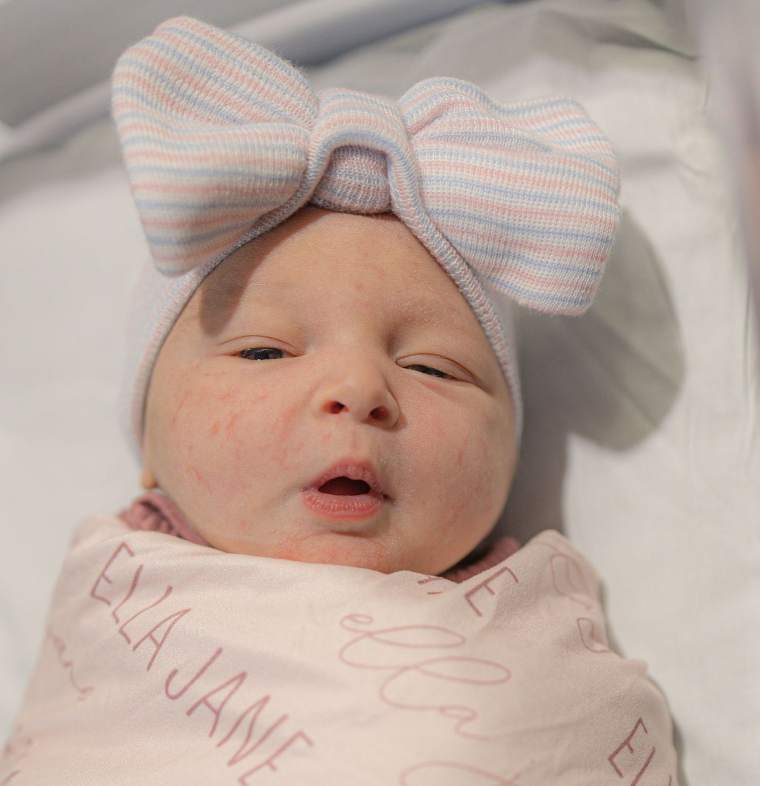 Newborn baby wearing a striped bow headband rests peacefully in white bedding.