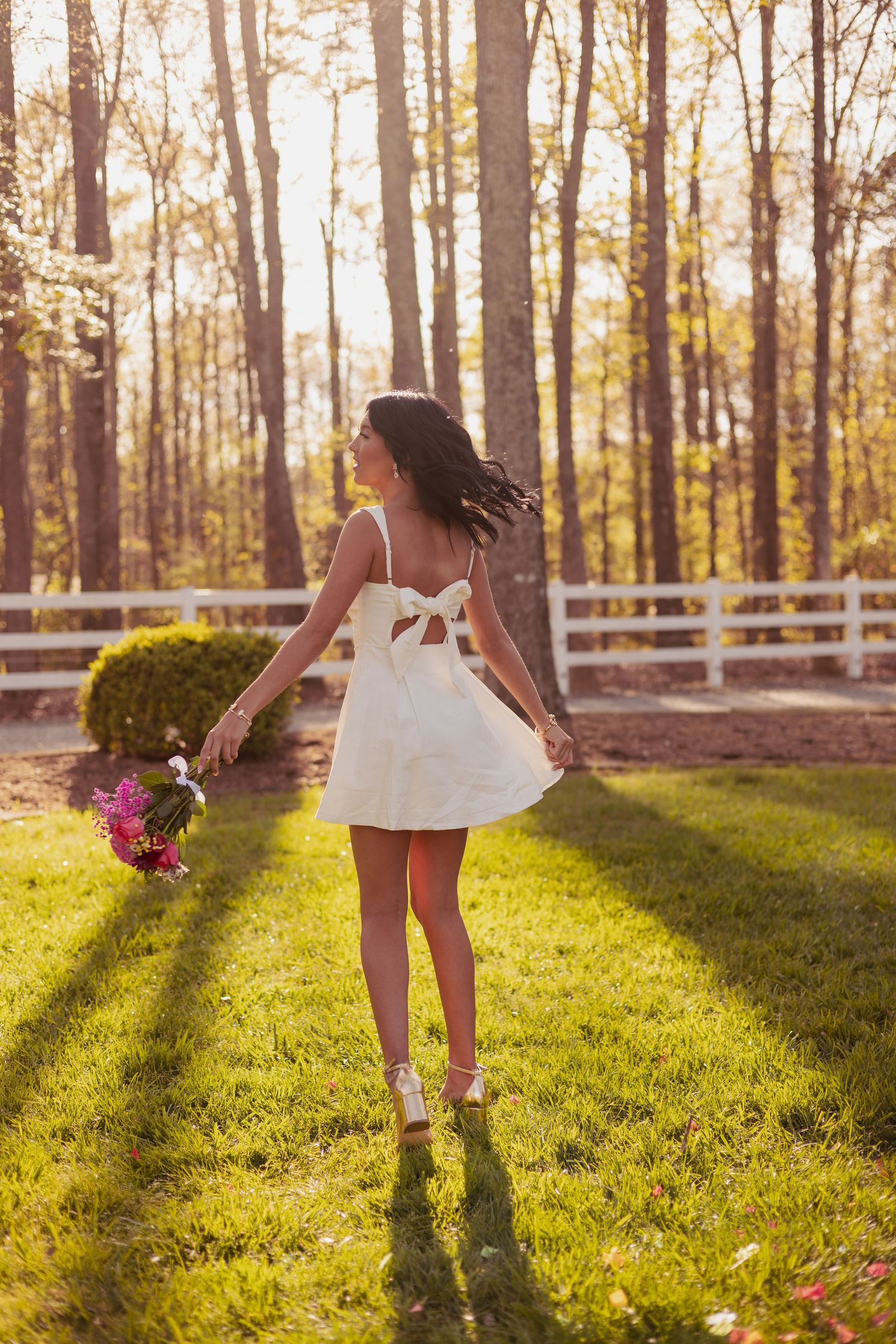 Young woman in white mini dress holding flowers, twirling in sunny green yard with white fence and tall trees.