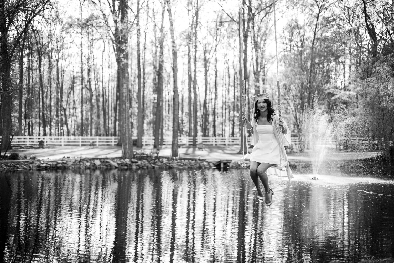 Black and white photo of a woman in a white dress sitting on a swing over a reflective pond in a forest.
