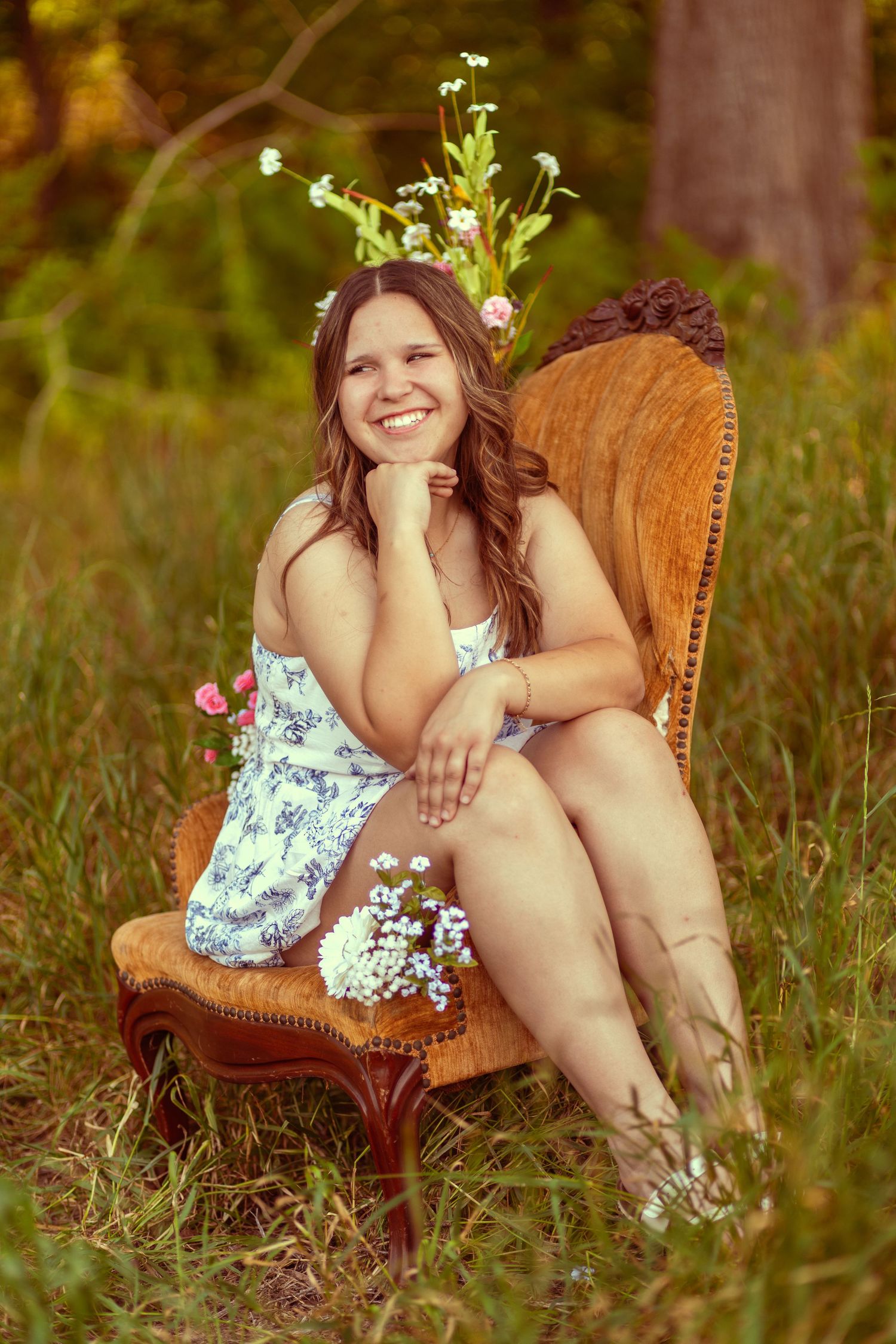 Smiling young woman in floral dress sitting in vintage chair outdoors with wildflowers, surrounded by lush green grass.