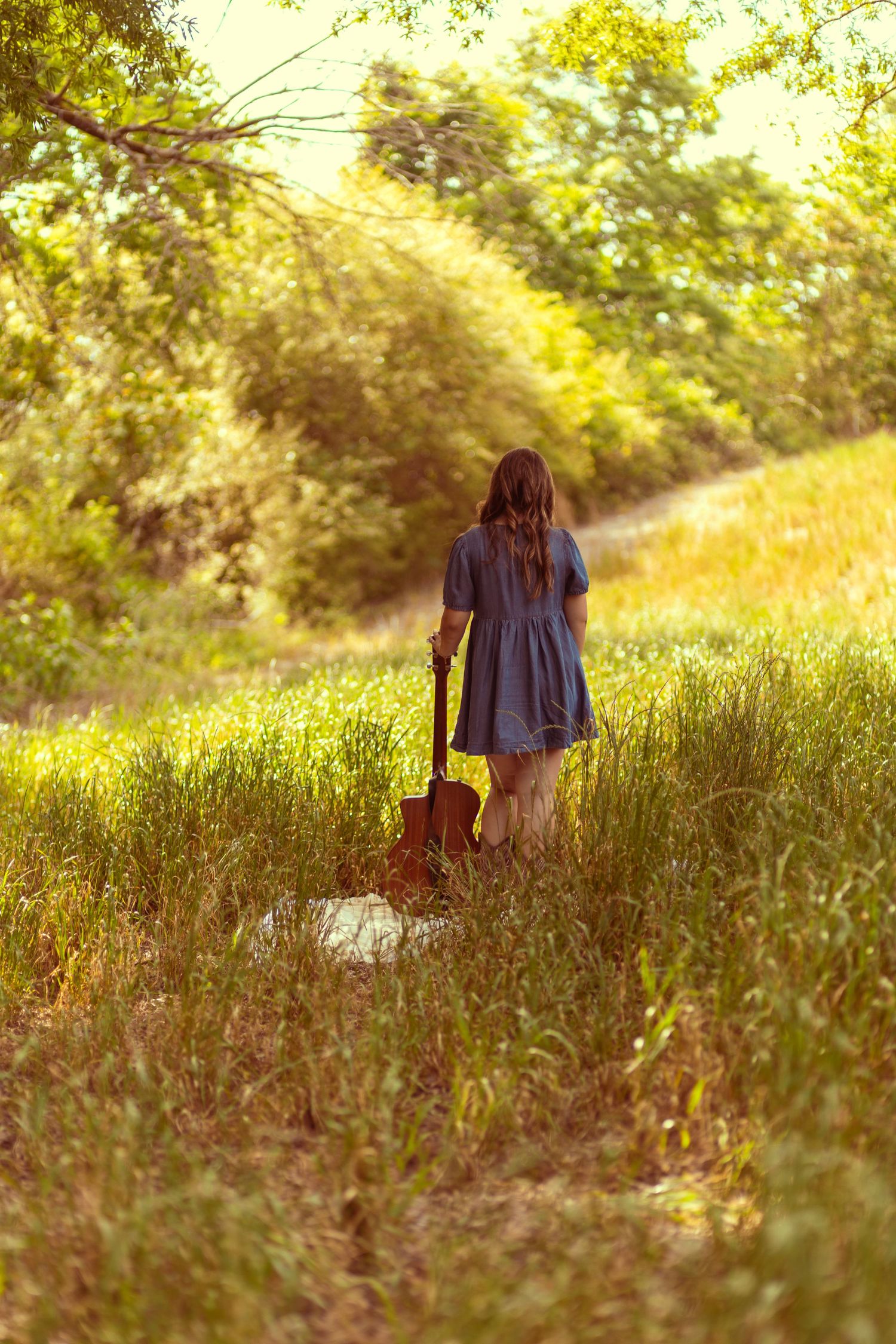Young woman in blue dress standing in sunny meadow holding acoustic guitar among tall green grass.