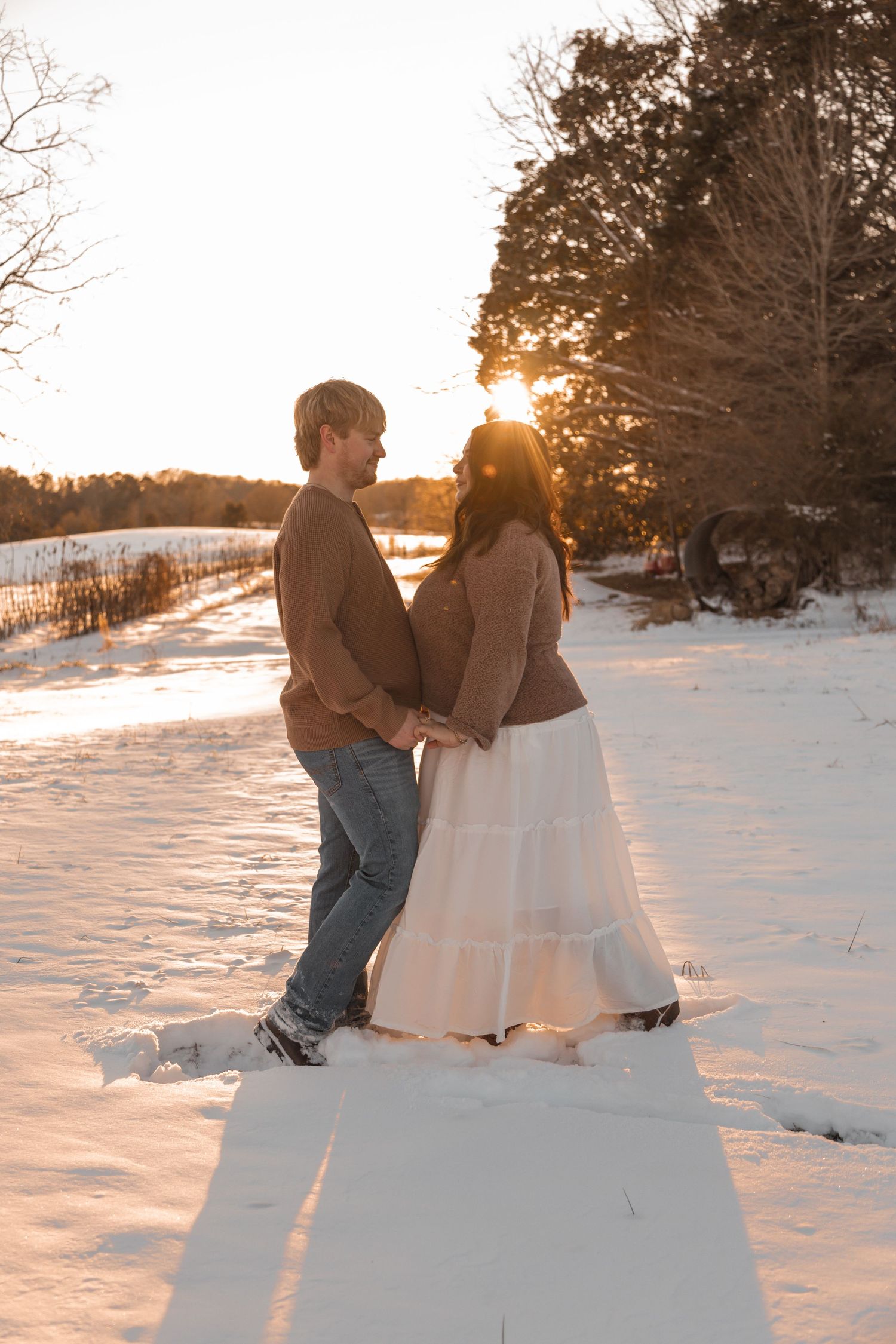Couple holding hands in snowy winter landscape at golden sunset, woman in white dress and man in brown sweater.