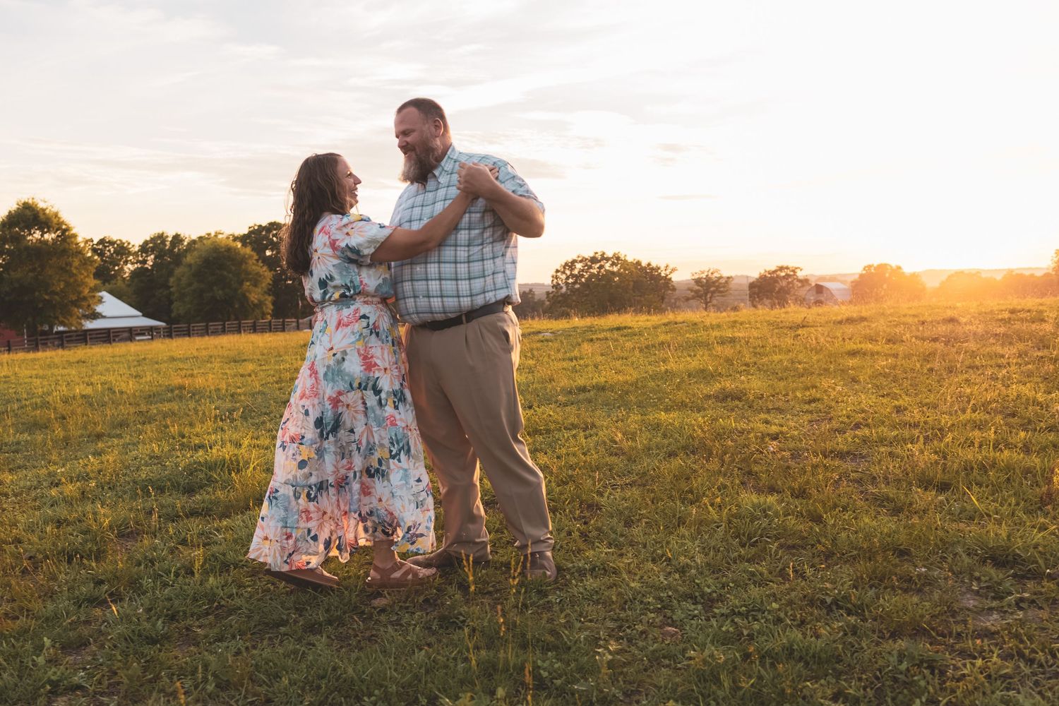 Couple dancing together in a sunlit green field at golden hour sunset, woman in floral dress, man in plaid shirt.