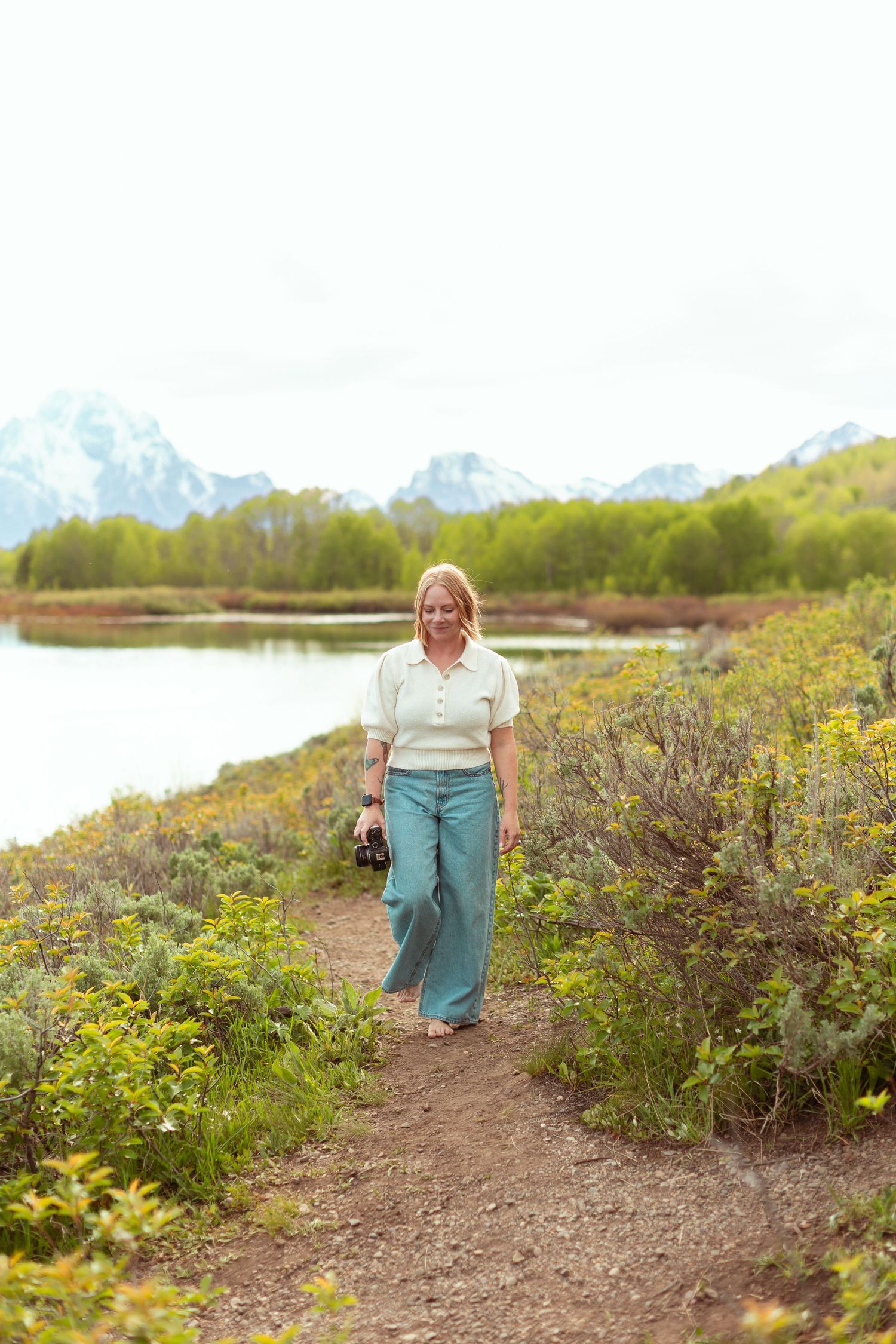 A smiling woman walks a scenic trail near a lake with snow-capped mountains in Grand Teton National Park.