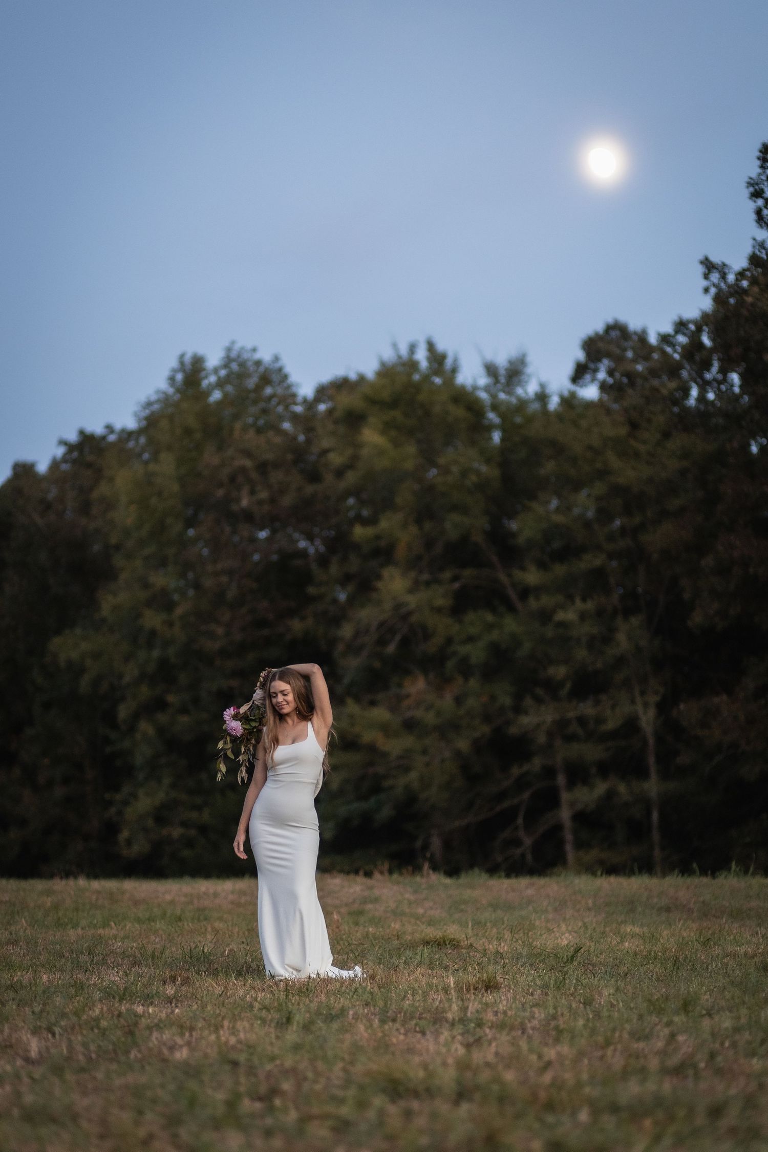 A figure in a white gown stands in a grassy field at dusk with trees and a bright moon in the background.