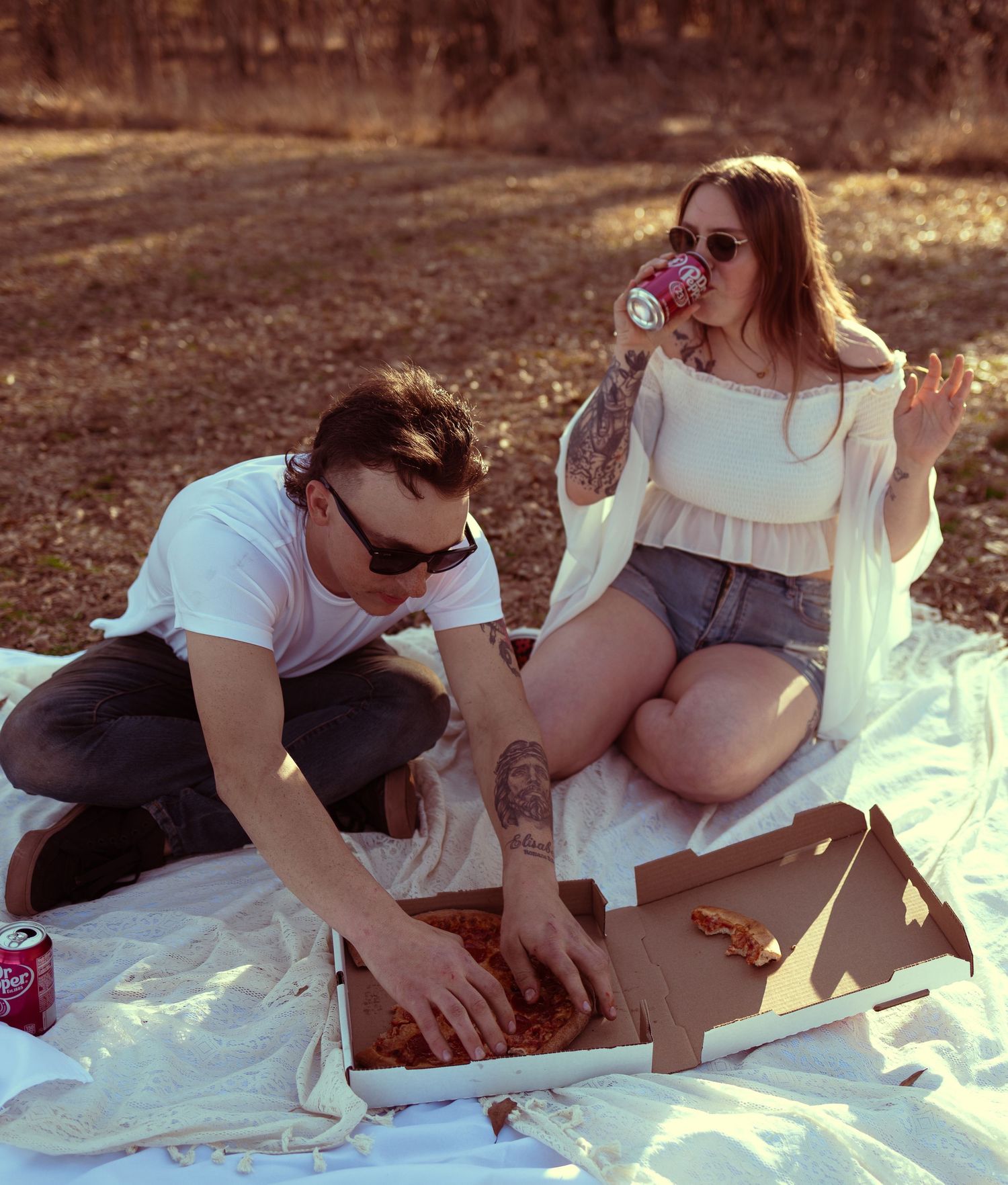 Couple enjoying a outdoor picnic with pizza and drinks on a white blanket in a sunny park setting.
