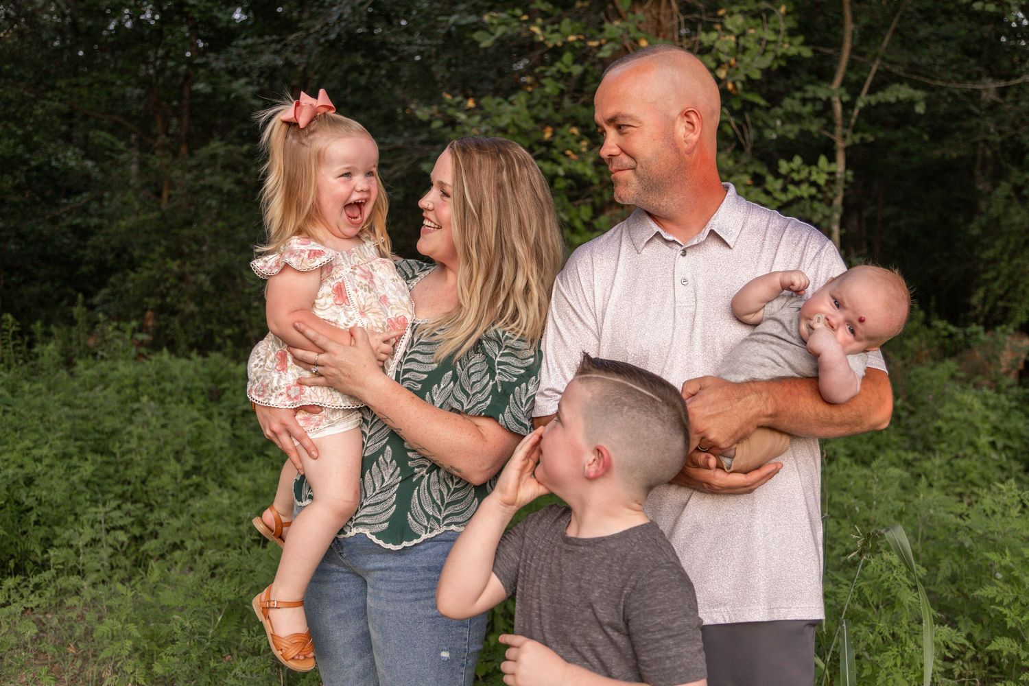 Family of four laughing and embracing together outdoors in a wooded setting while wearing casual summer attire.