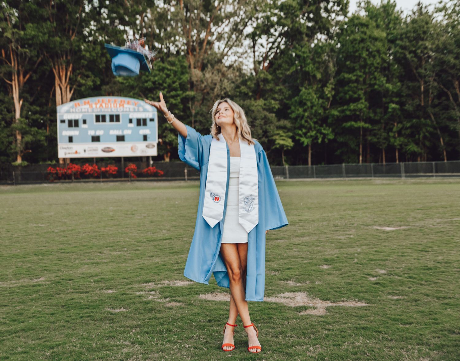 Graduate in blue cap and gown standing on a sports field celebrating with pom poms.
