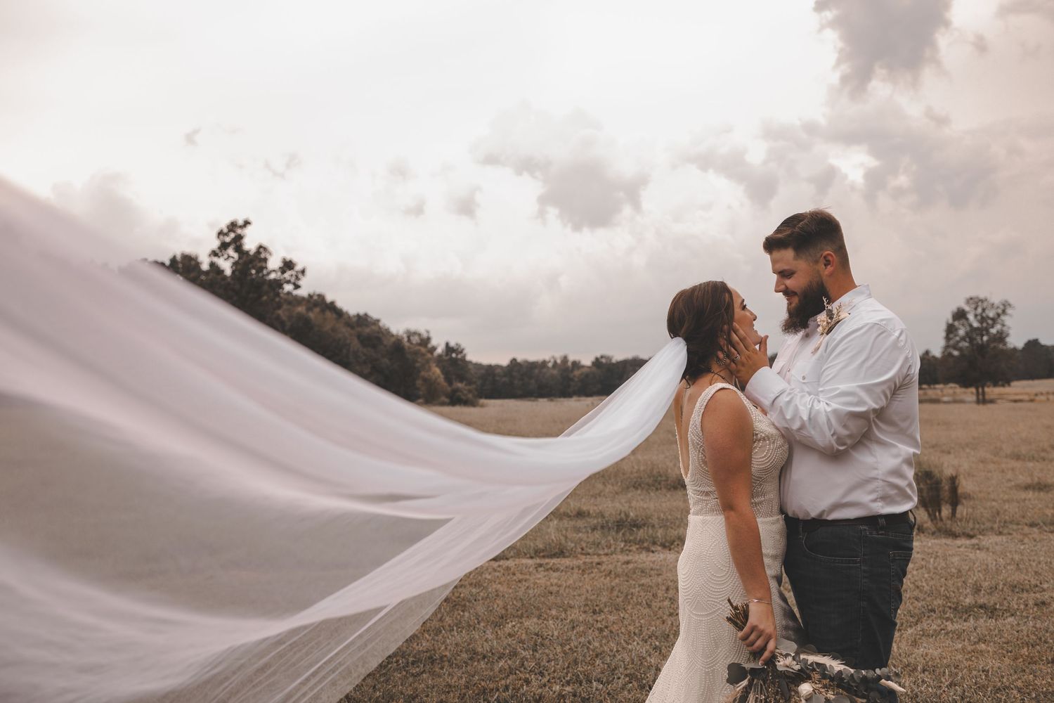A couple embraces in a romantic outdoor setting with a flowing white veil catching the wind during a sunset photoshoot.