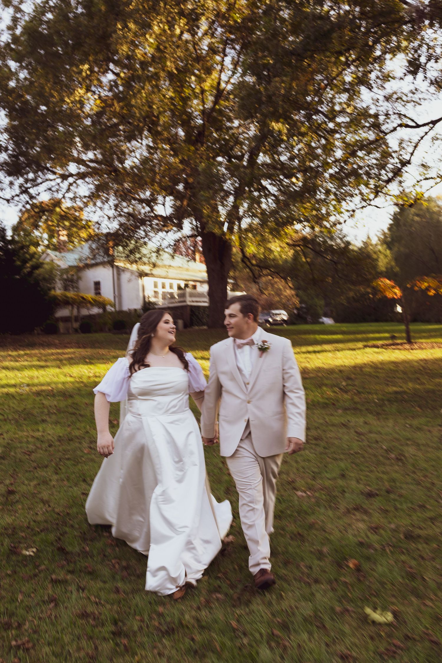 A couple in wedding attire walks together on a grassy lawn during golden hour with trees and a building in the background.