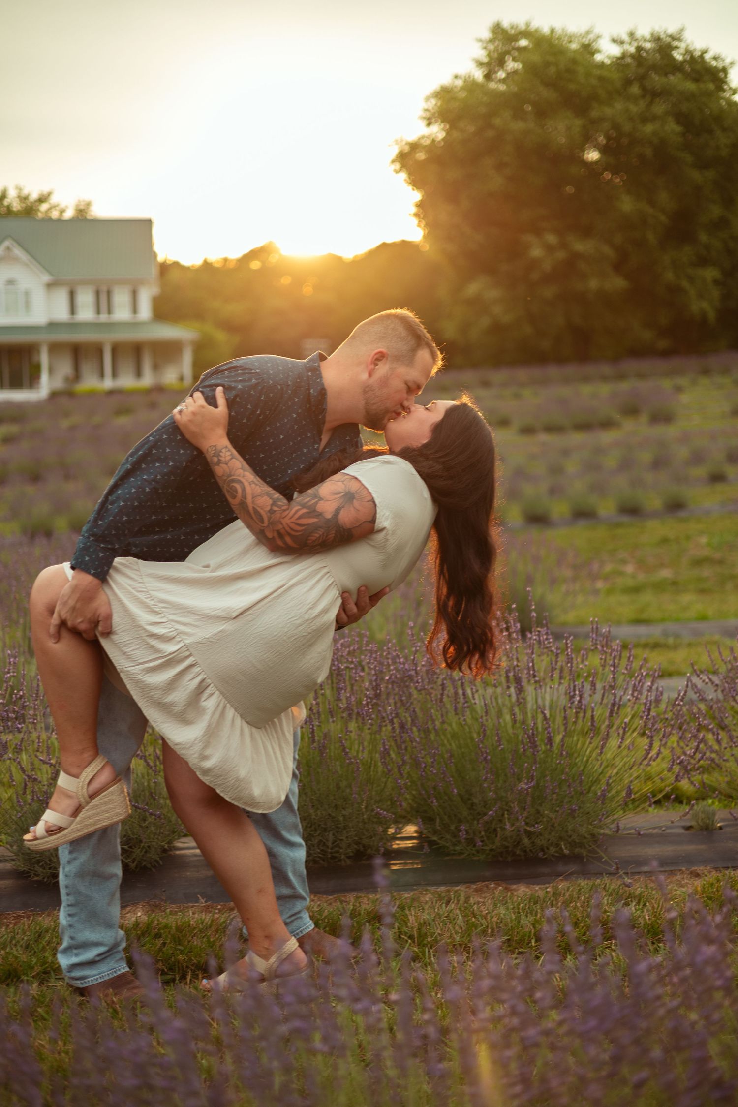 Couple sharing a romantic dip kiss in a lavender field at golden sunset, with a white farmhouse in the background.