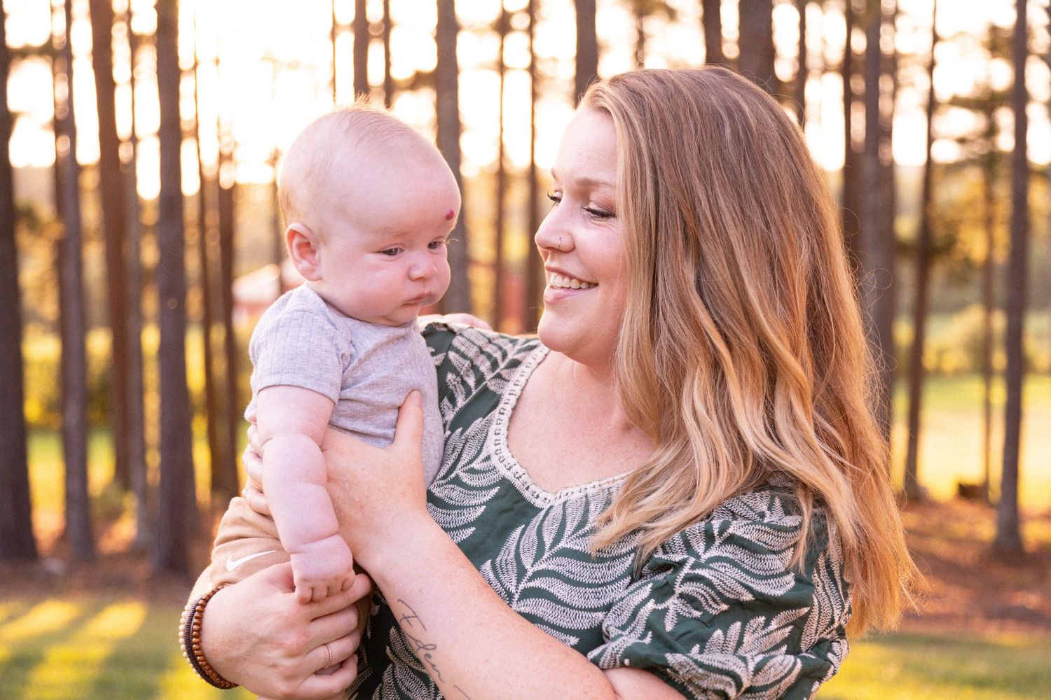 Mother holding baby outdoors at sunset with glowing forest light and pine trees in background.