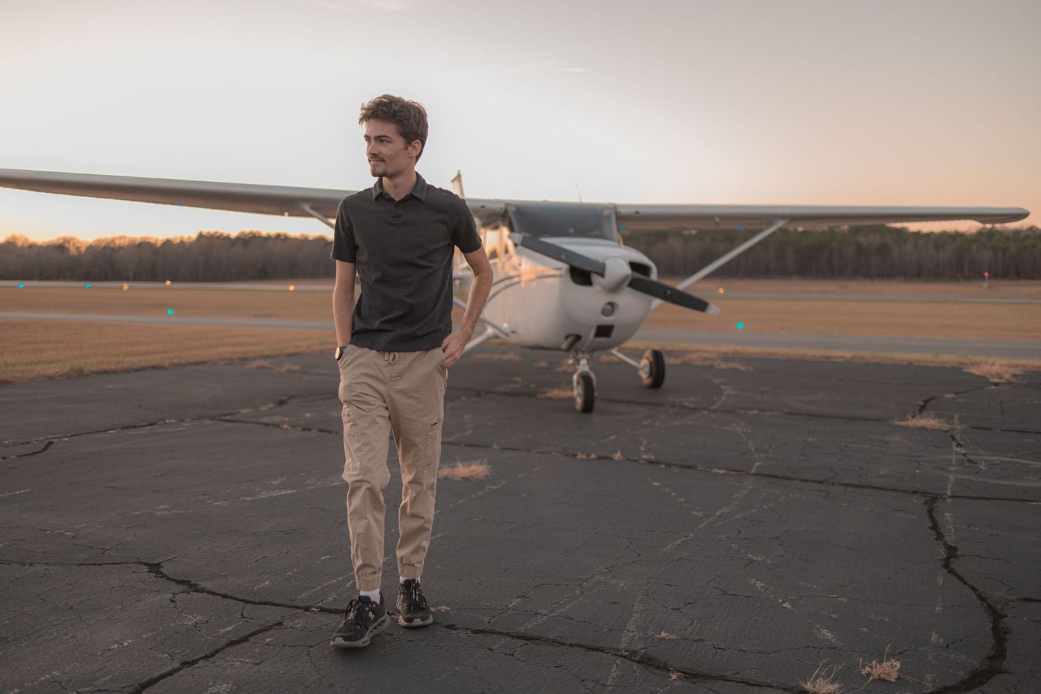 A person stands near a small private airplane on a runway during sunset with warm golden lighting.