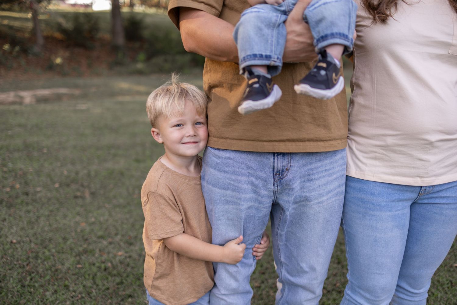 A toddler in a brown shirt hugs someone wearing blue jeans while standing on grass outdoors.