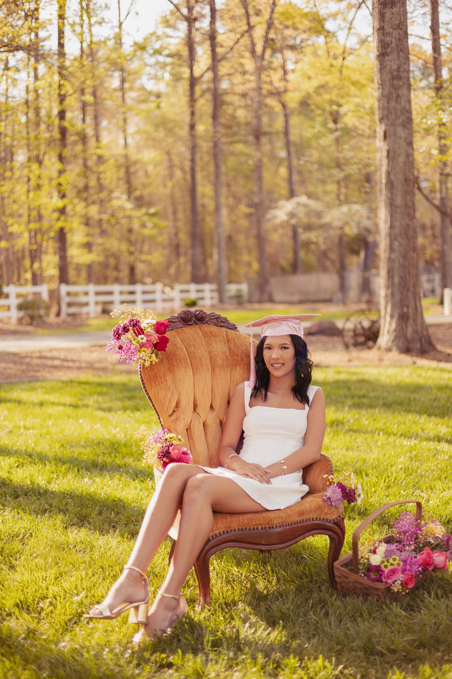 Young woman in white dress and pink graduation cap sits in ornate chair outdoors, surrounded by pink flowers.