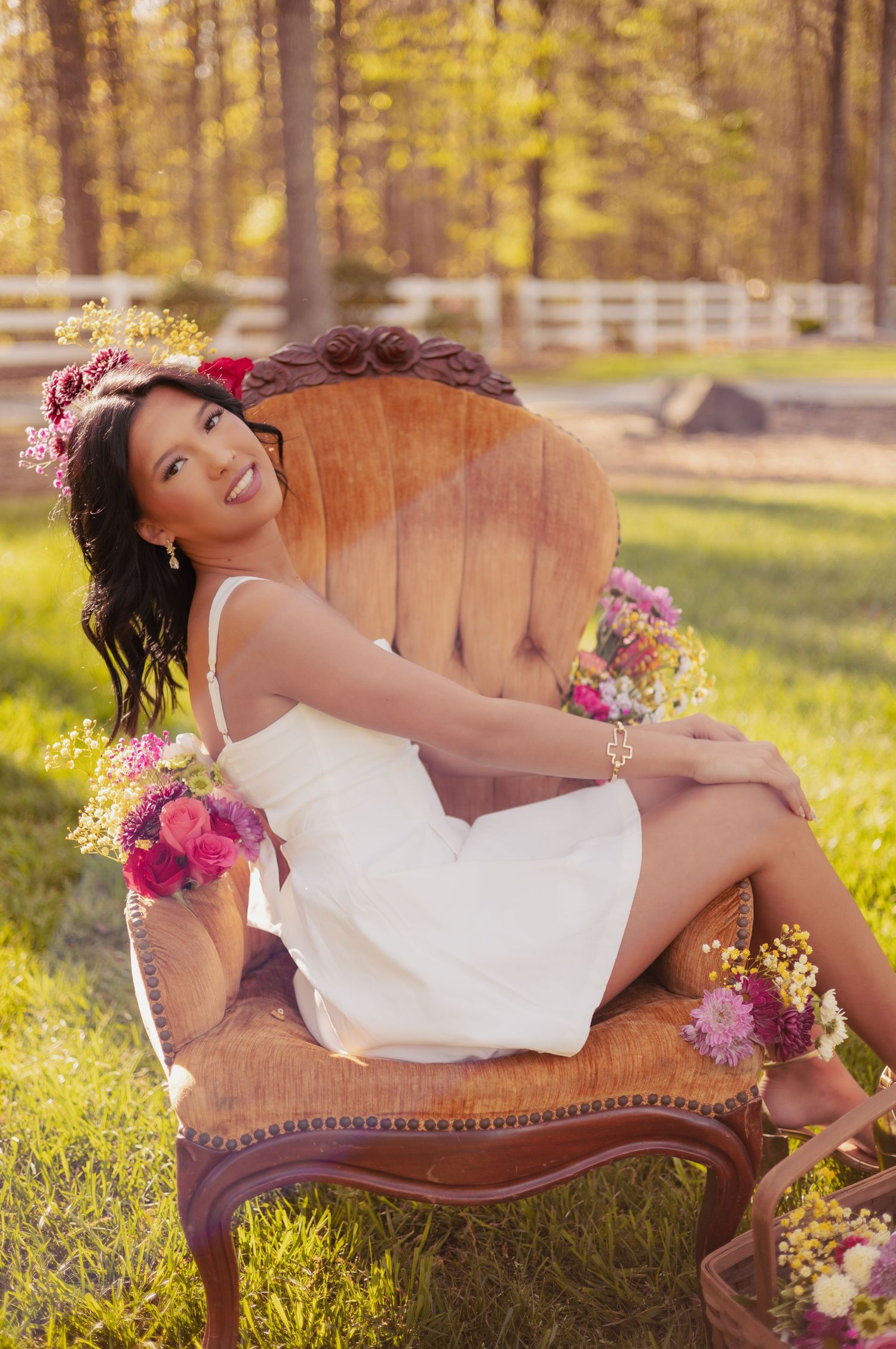Smiling woman in white dress with floral crown sits on vintage wooden chair outdoors surrounded by colorful flowers.