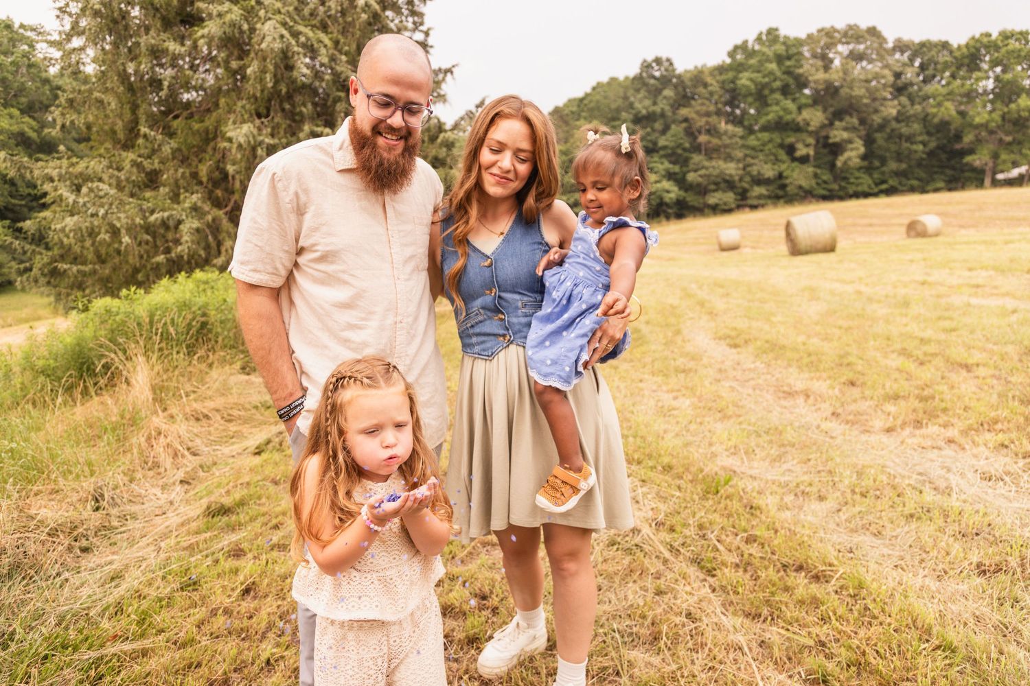 Family laughs and plays together in a sun-drenched summer field with golden grass and rolling hills.