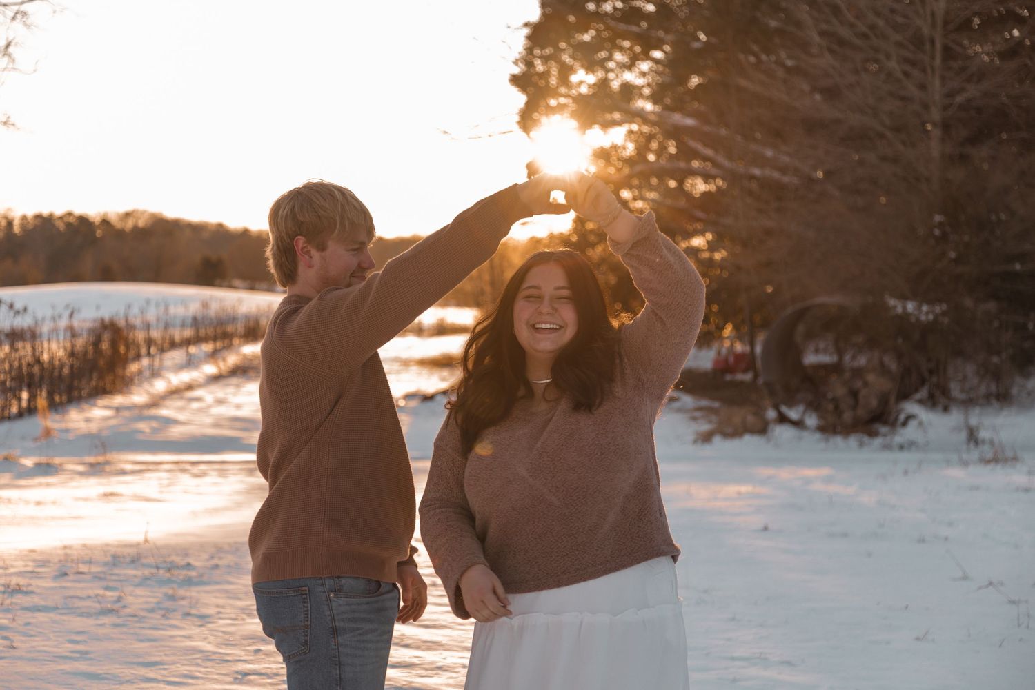 Couple dancing joyfully outdoors in snow during a warm golden sunset winter engagement session.