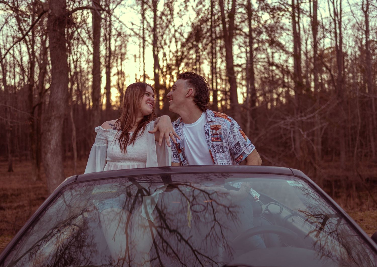 Couple standing through car sunroof smiling at each other surrounded by tall trees at sunset.