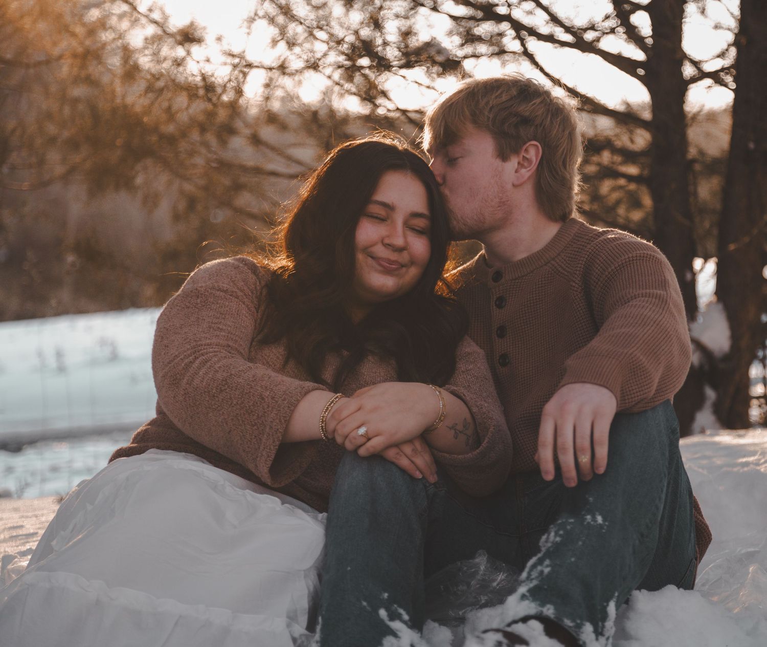 Couple sitting together in snow at golden hour, woman smiling as man kisses her cheek, winter outdoor romance.