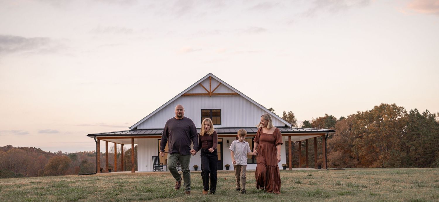Rural landscape at sunset with a white farmhouse and barn visible in the distance as people walk across the field.