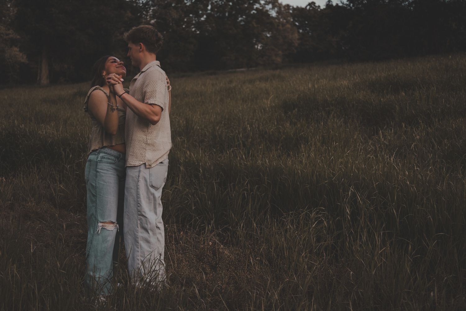 A couple embraces in a dimly lit field at dusk wearing casual attire.