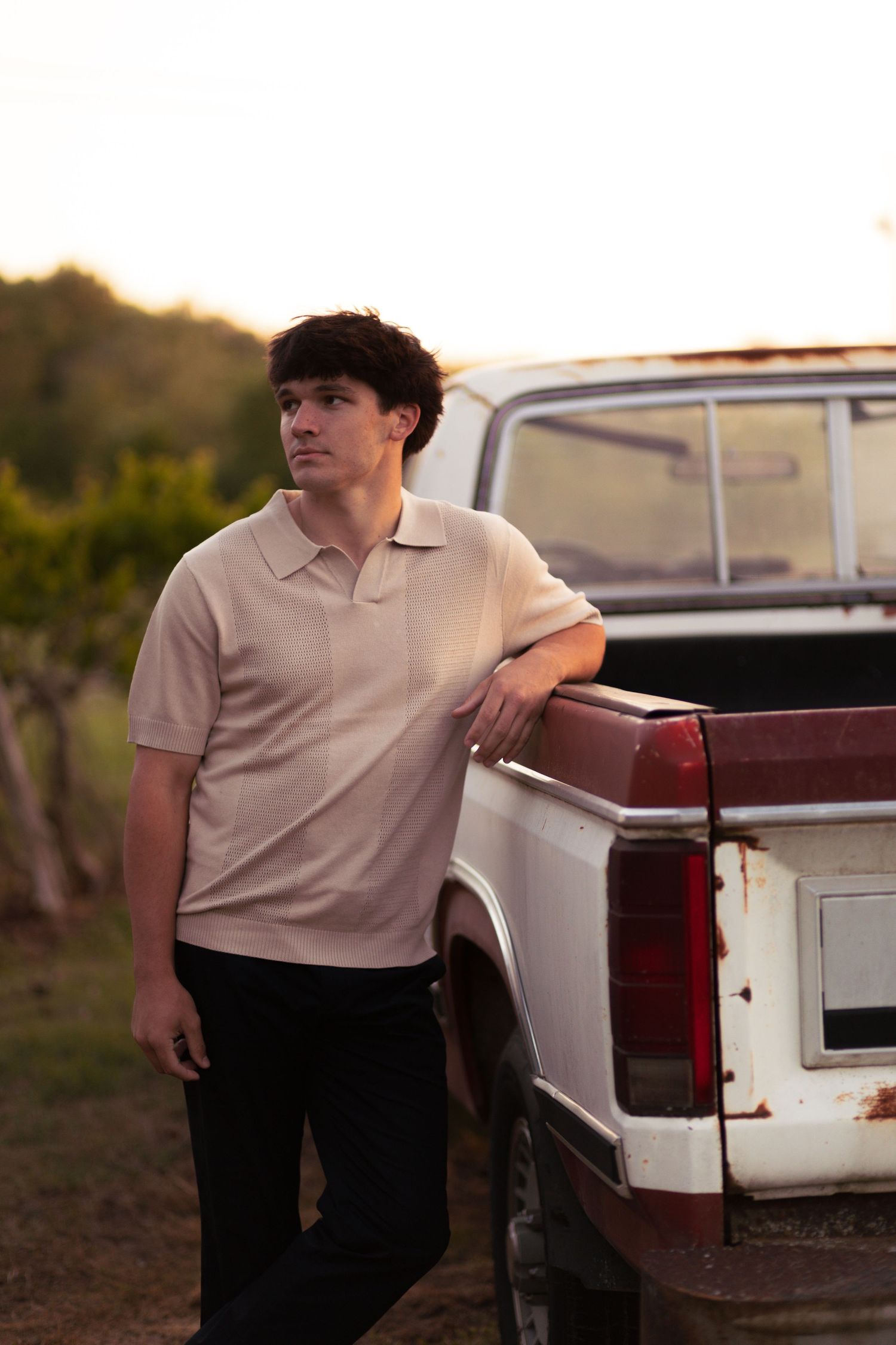 Young man in beige polo shirt leaning on vintage rusty pickup truck at golden hour in rural vineyard.
