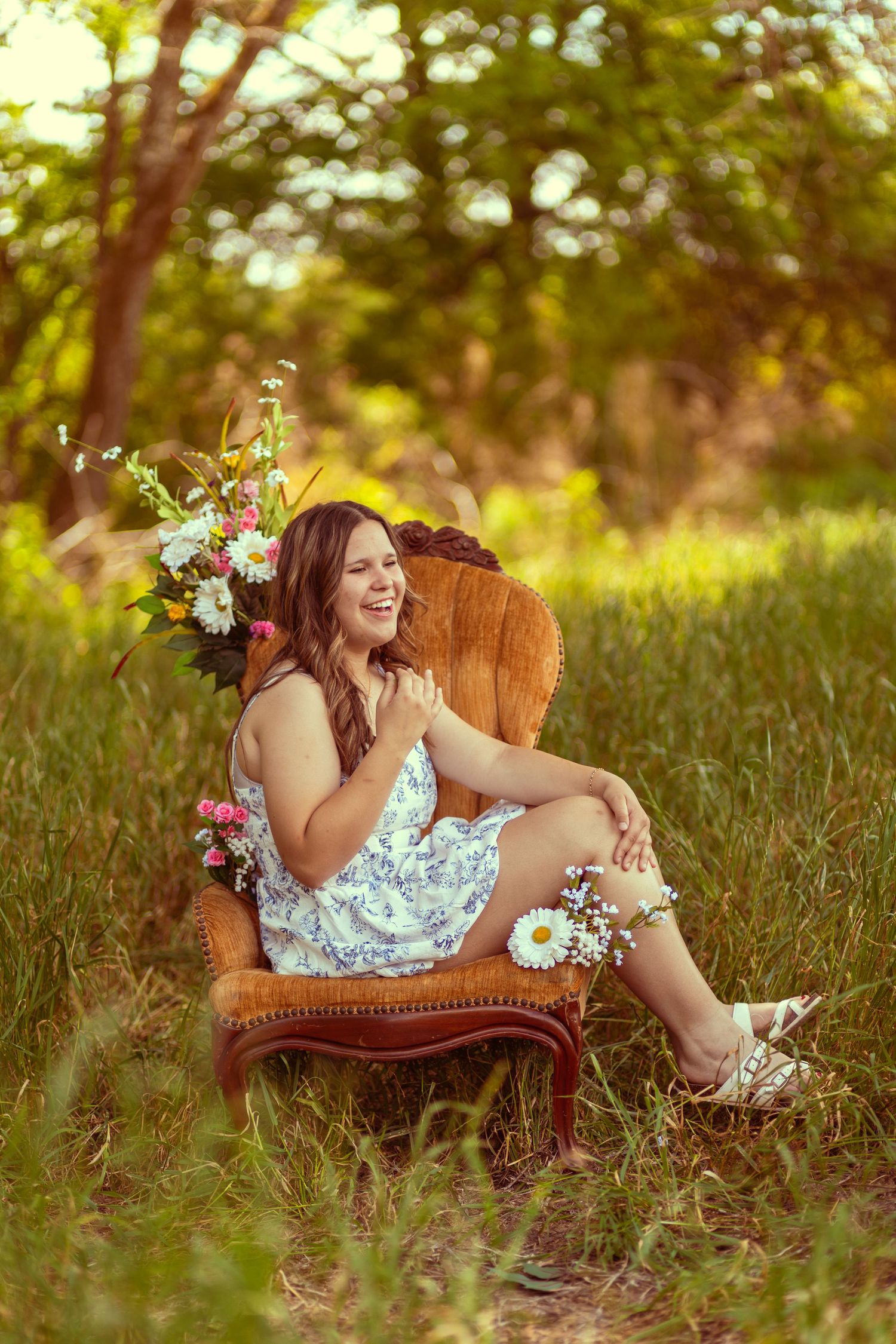 Young woman in floral dress laughing on vintage chair outdoors, surrounded by wildflowers and lush green trees.