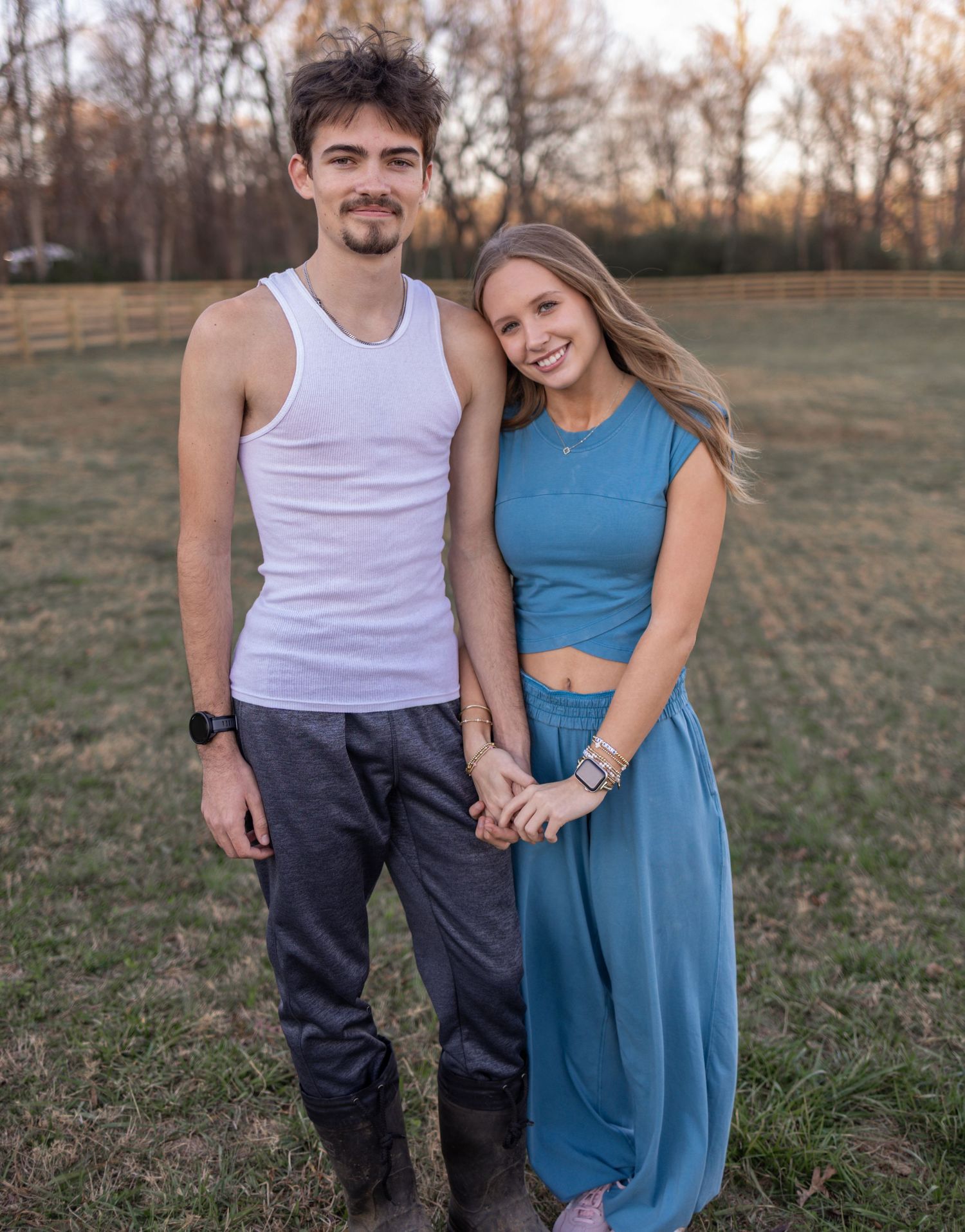 A couple stands together in an outdoor field during golden hour wearing a white tank top and blue dress.