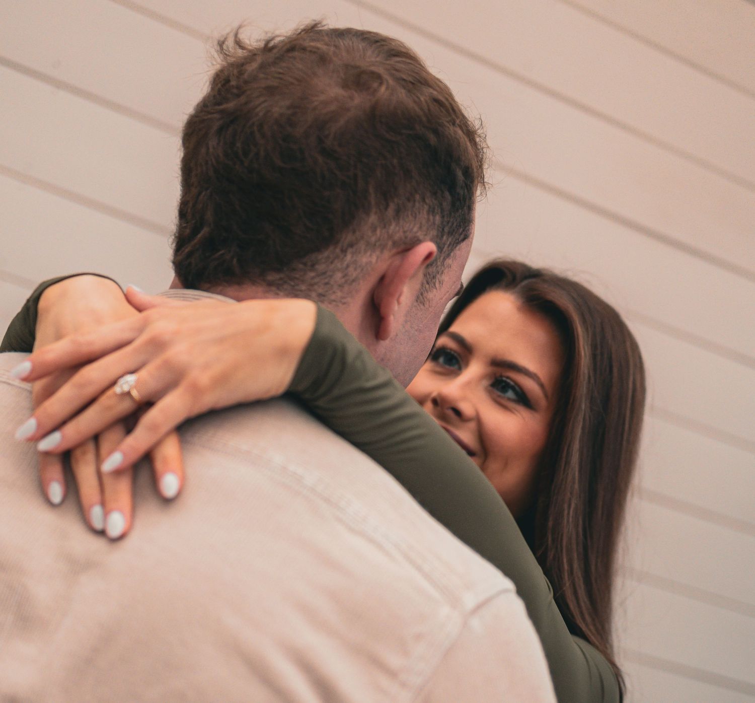 A couple in formal attire embraces in a tender moment against a beige wall.