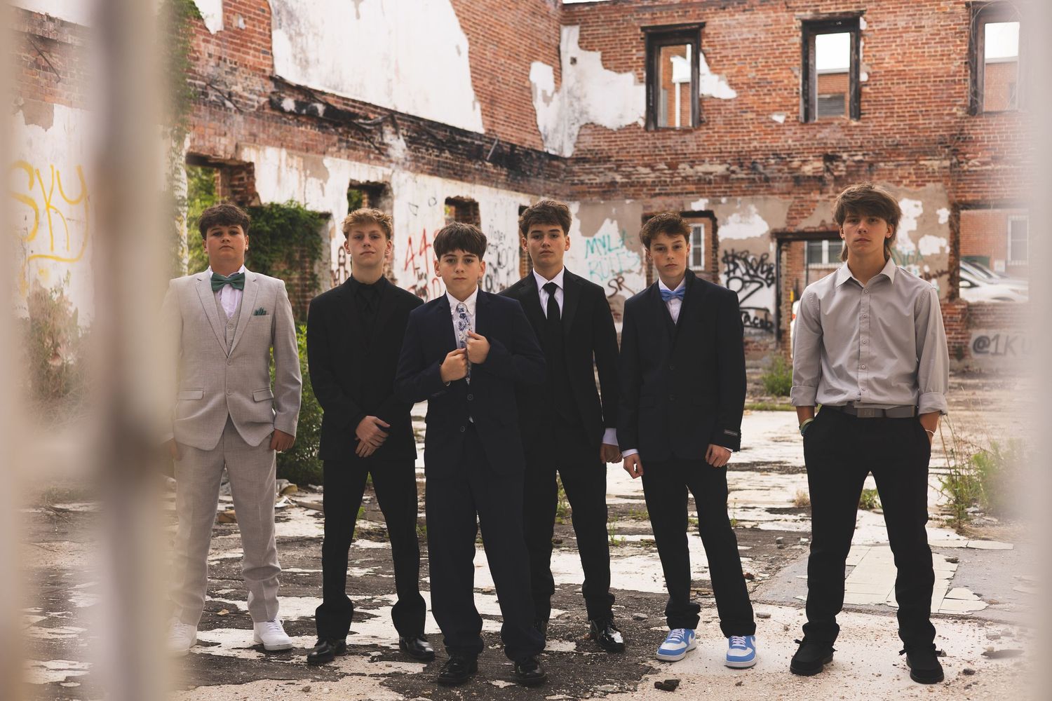 Group of teenage boys in formal suits posing in front of a rustic brick building for prom photos.