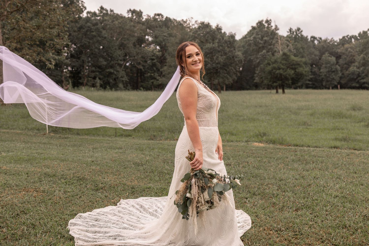 A bride in a flowing white wedding dress stands in a grassy field as her veil billows dramatically in the wind.