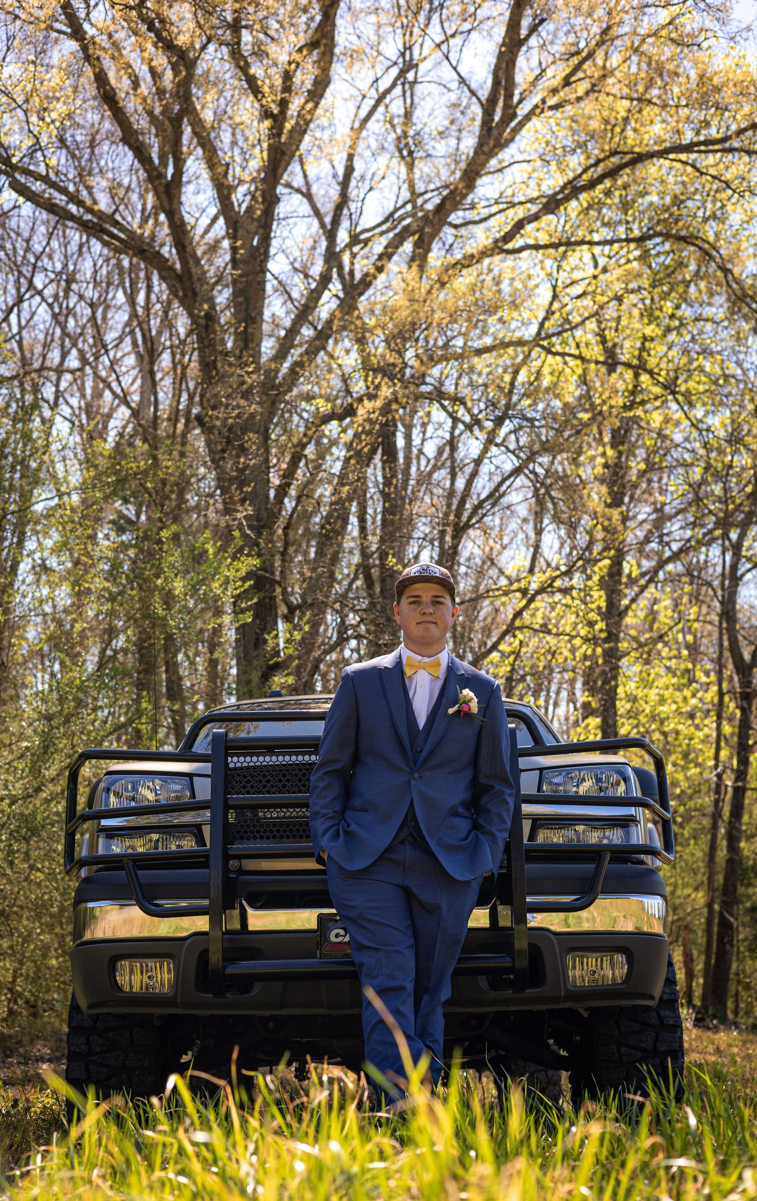 Young man in blue suit with boutonniere leans against black truck bumper surrounded by spring trees.