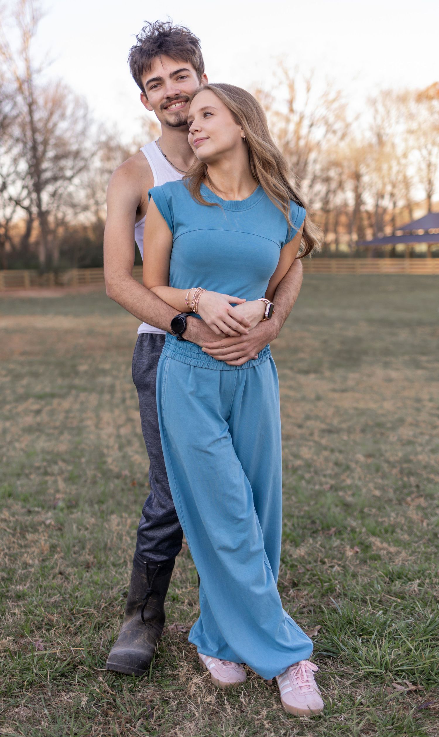 A couple embraces in a casual outdoor setting at sunset, with her wearing a blue outfit and him in a sleeveless top.