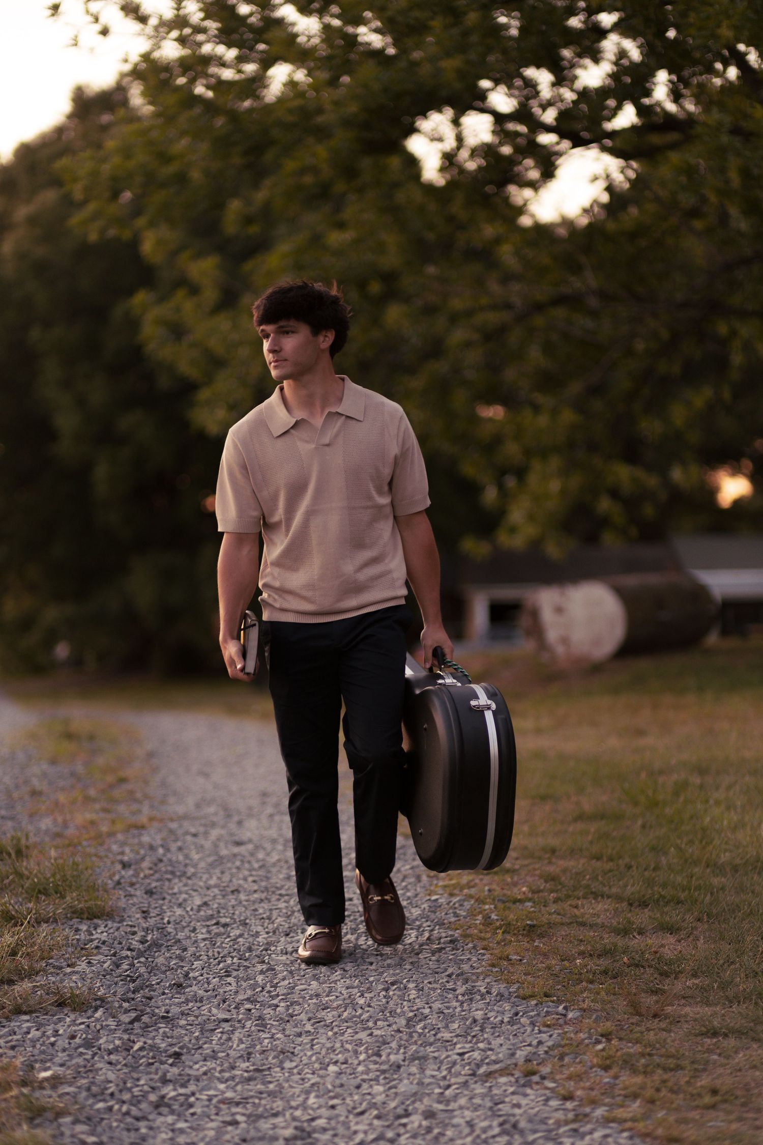 Young man in beige polo walks gravel path at sunset carrying a guitar case amid lush green trees.