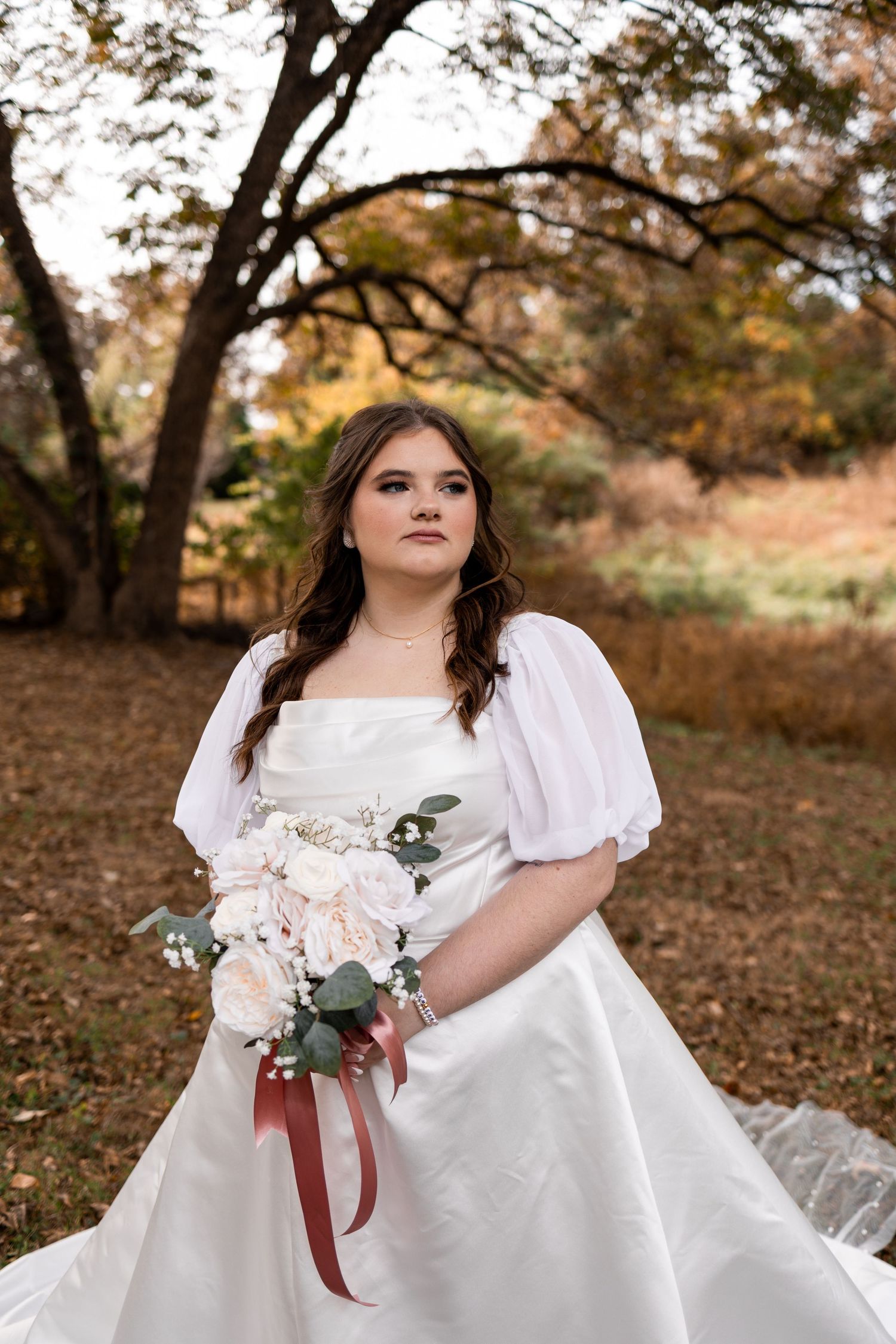 Bride in white gown with puff sleeves holds bouquet of blush roses outdoors in autumn setting.