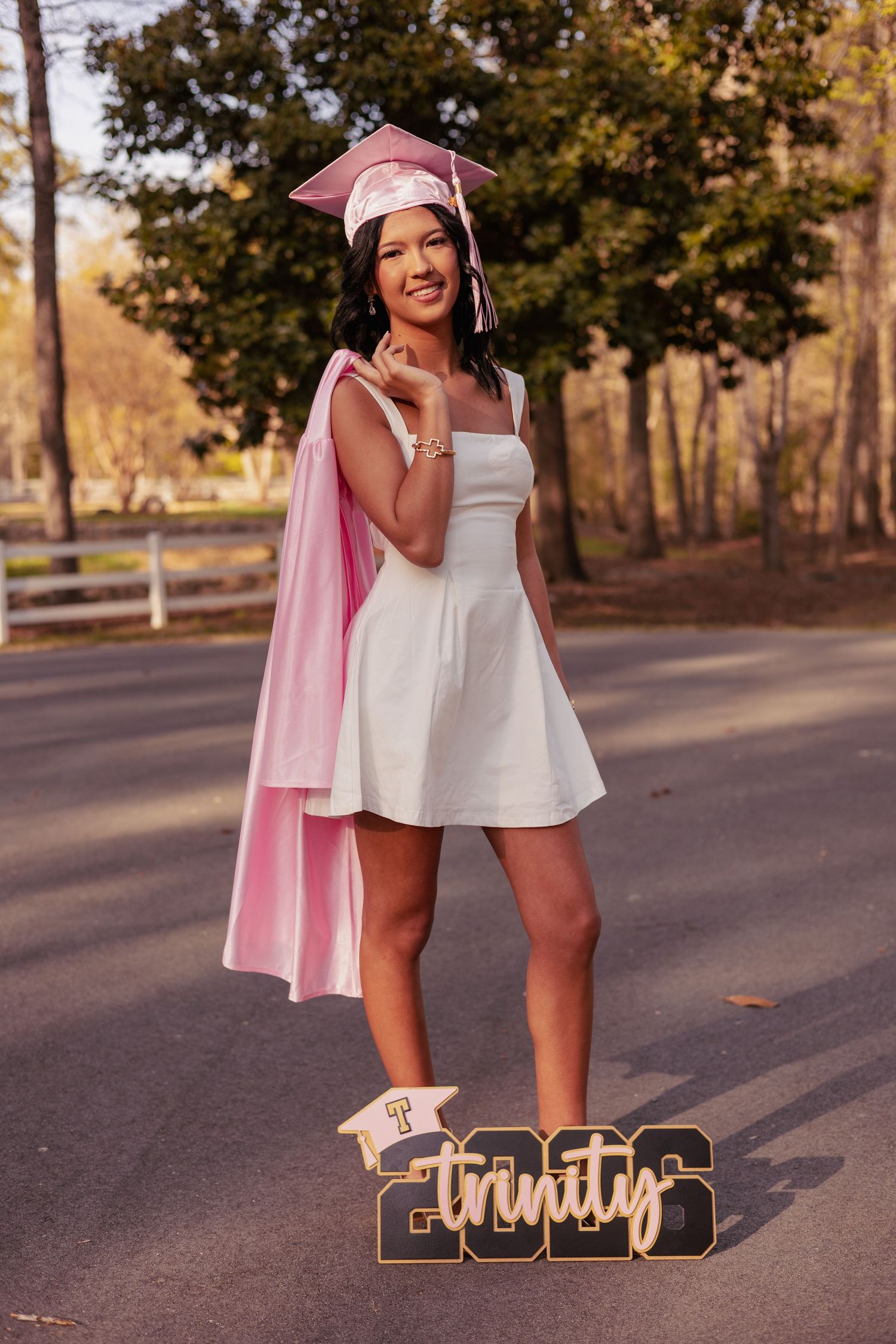 Young woman in white dress and pink graduation cap and gown poses on road with Trinity 2026 sign.