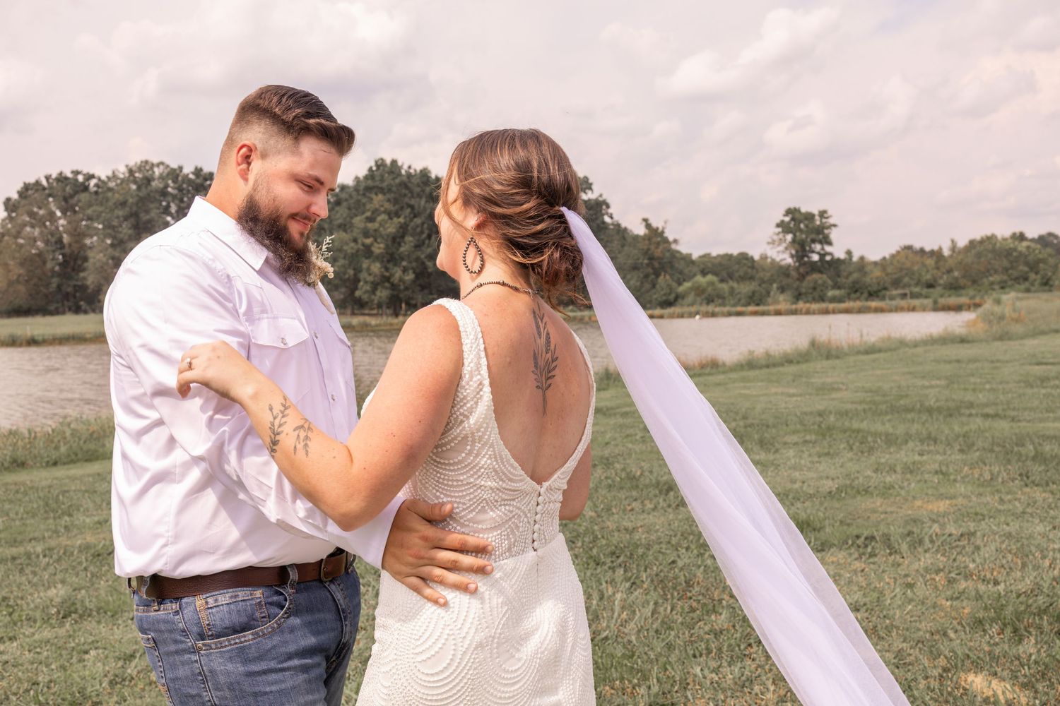 Romantic wedding couple embraces in a sunlit meadow with a flowing white veil caught in the breeze.