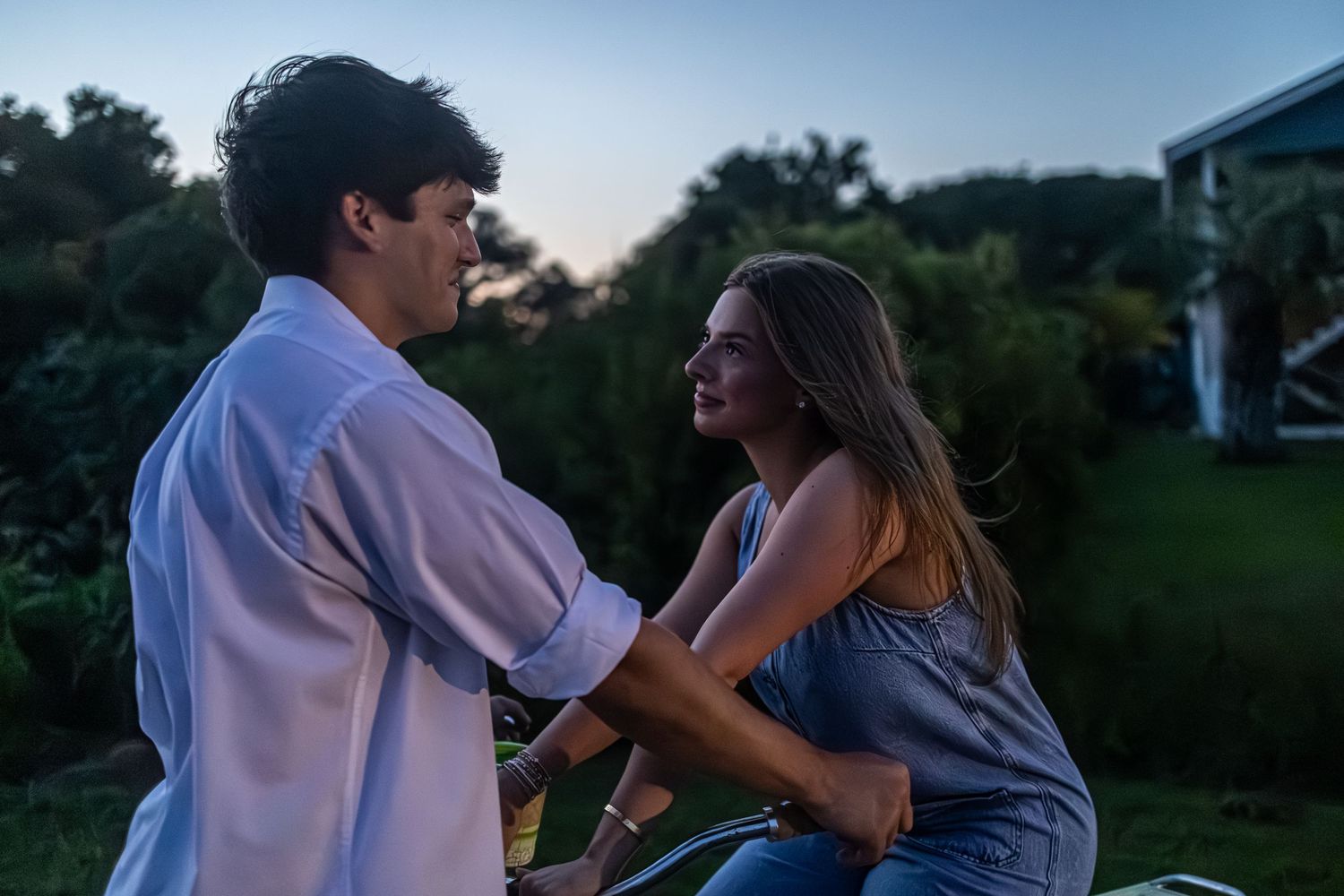 A young couple shares a romantic moment outdoors at dusk, surrounded by lush greenery.