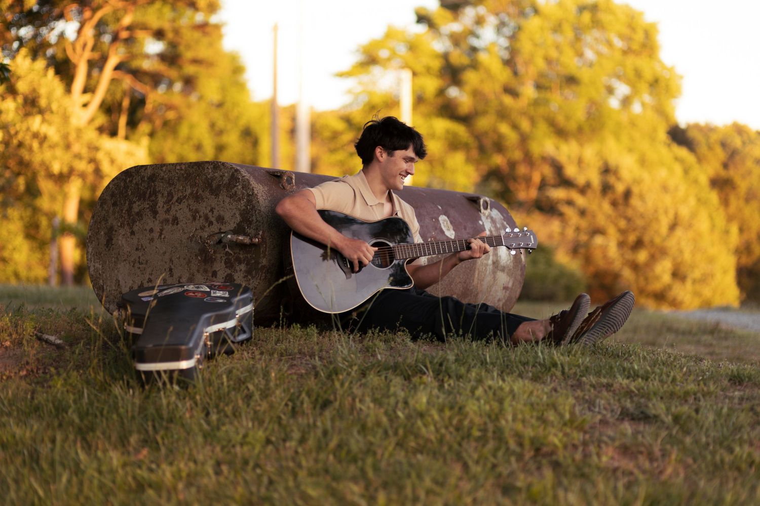 Young musician playing acoustic guitar outdoors on grass at golden hour, leaning against a large hay bale.
