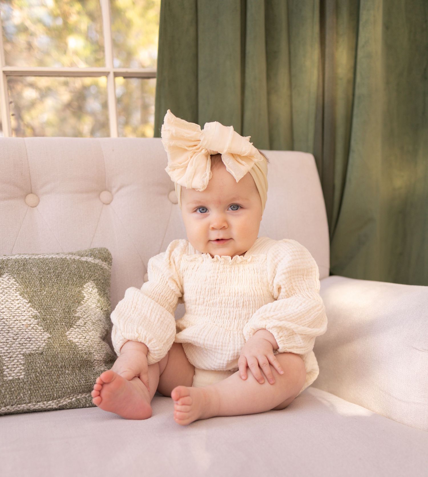 A baby wearing a pink bow and romper lies on a white couch in a softly lit room with green curtains.