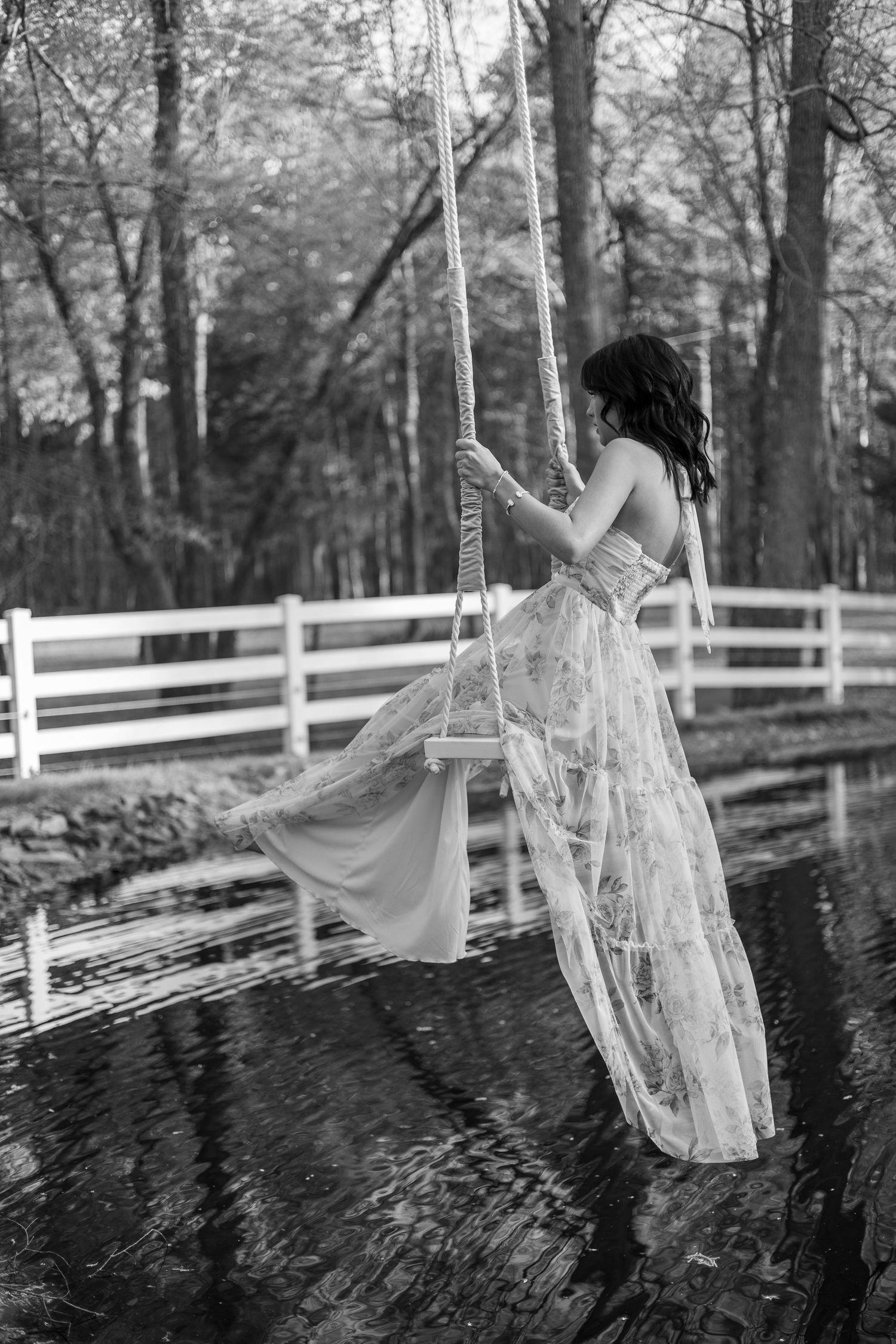 Black and white photo of a woman in a lace gown sitting on a rope swing near a reflective pond.