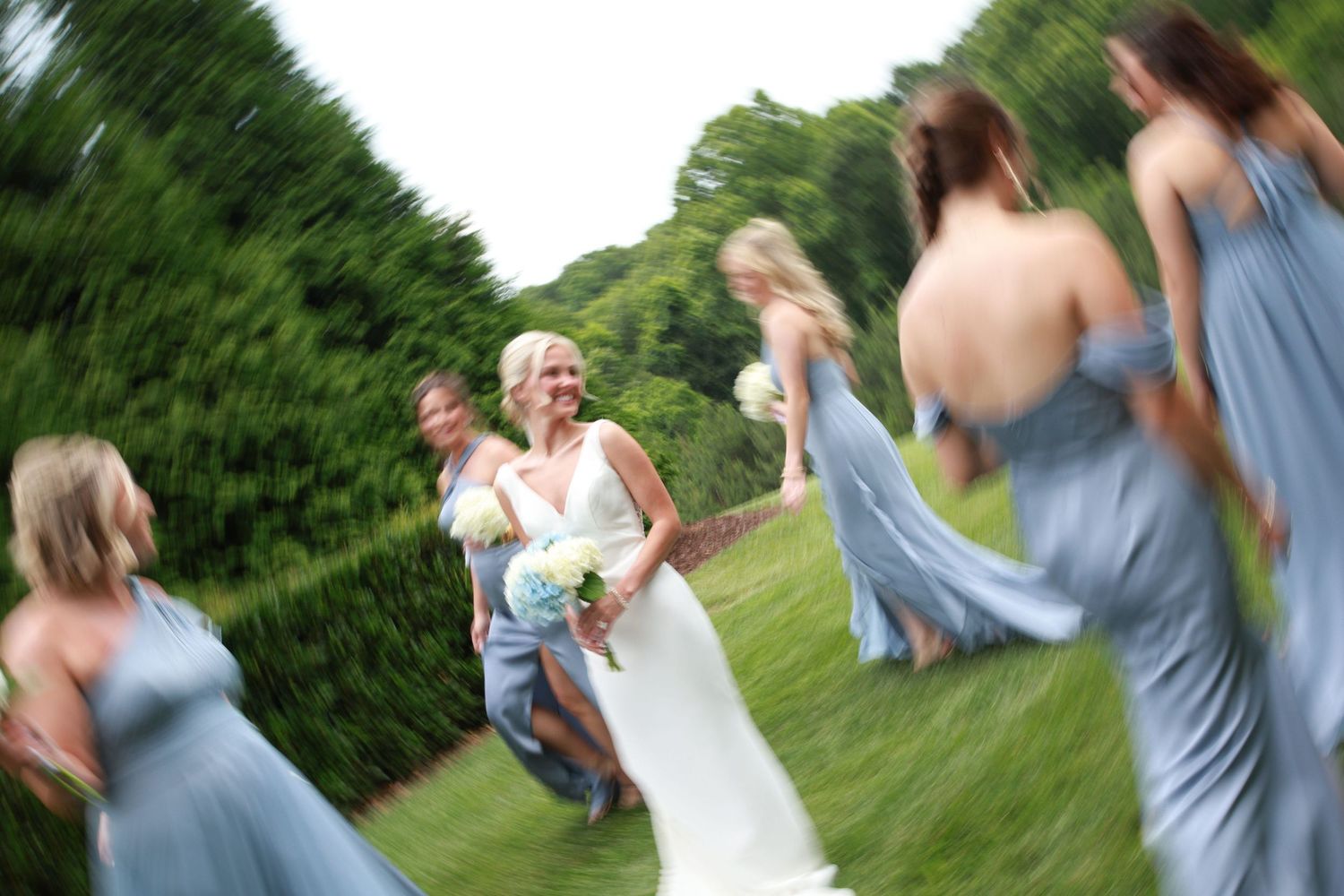 Wedding party celebrating outdoors with bridesmaids in blue dresses dancing in motion-blurred shot.