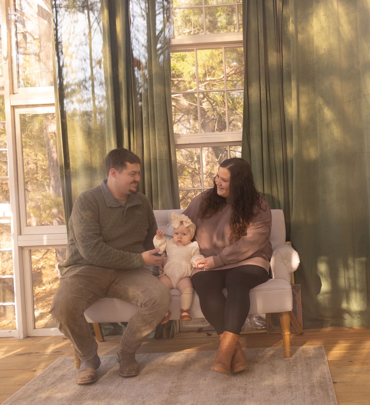 A family sits together in a sunlit room with large windows and wooden floors during a tender moment.