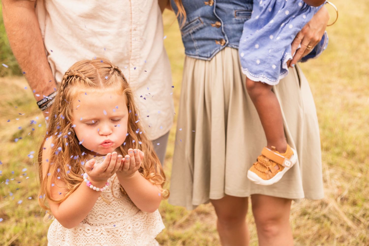 Child in a lace dress blowing on a dandelion flower in a sun-lit field during golden hour.