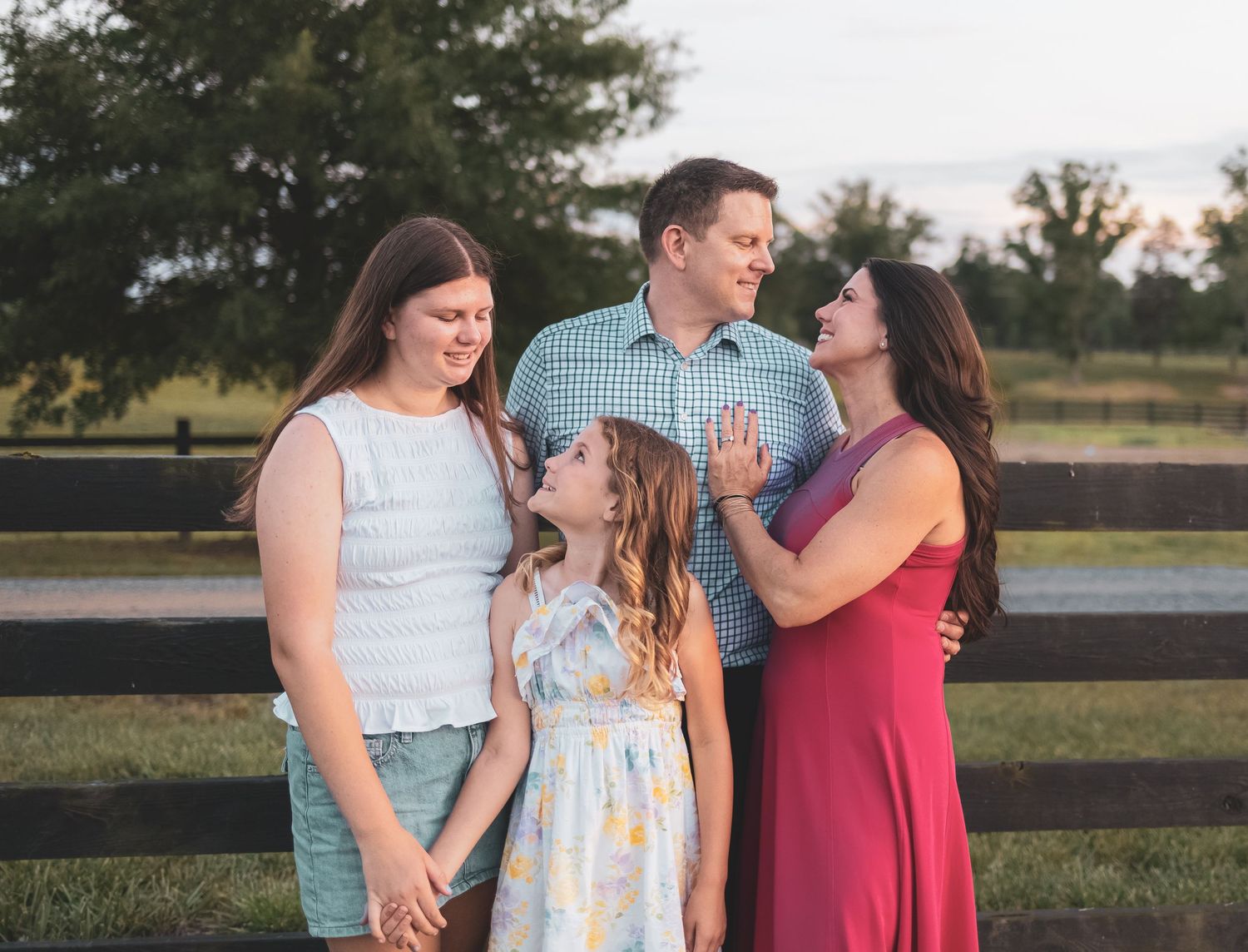 Family of four smiling and embracing outdoors near a wooden fence at golden hour.