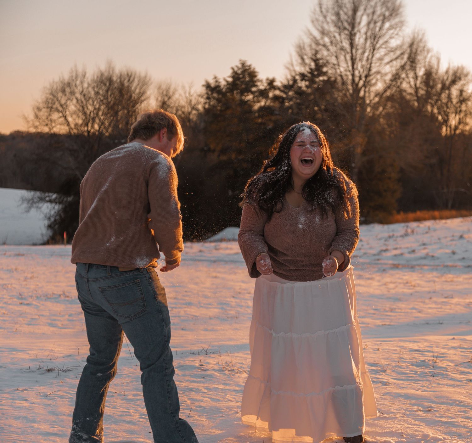 Couple laughing and playing in snow at golden hour sunset with bare trees in winter background.