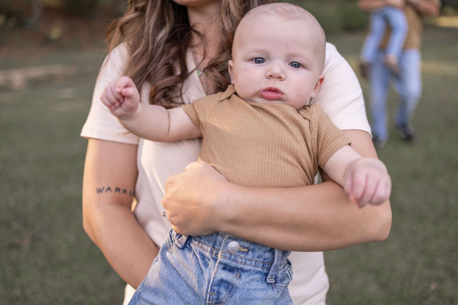 A baby wearing a brown sweater and jeans is held while standing in a grassy outdoor setting.