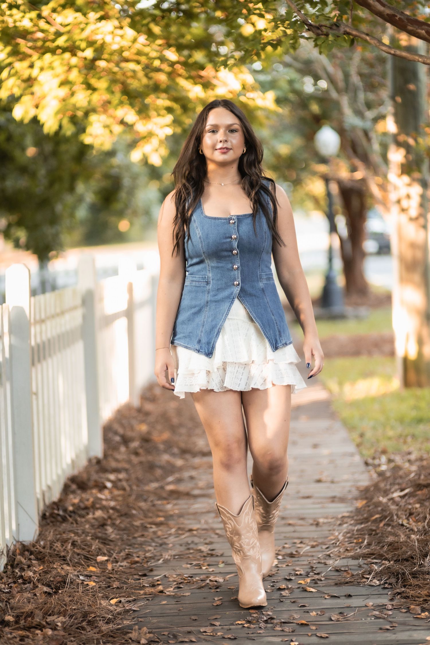 A person in a denim vest and white dress walks along a picturesque path lined with white picket fences and autumn leaves.